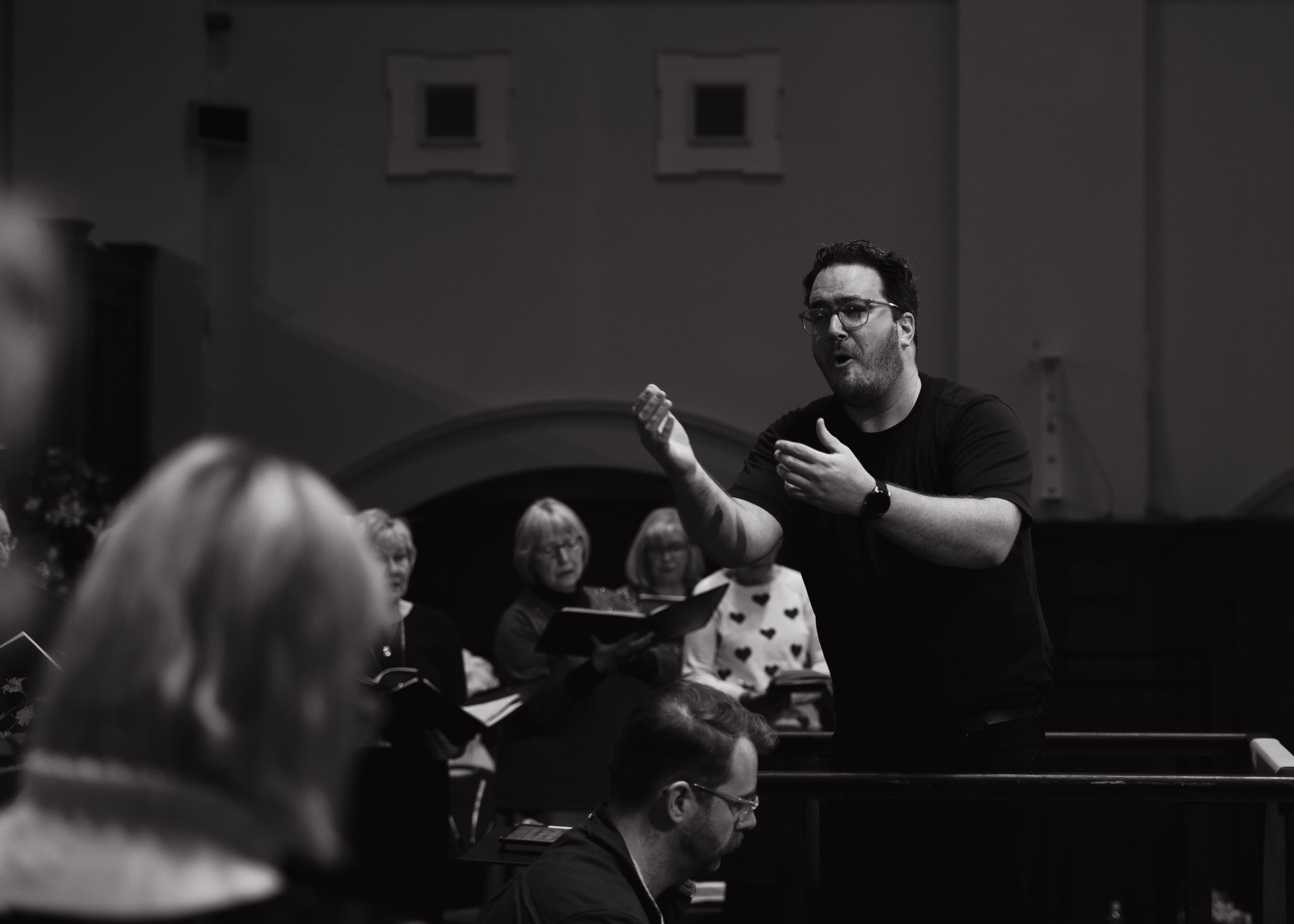 A conductor singing and gesturing while directing a seated choir, black and white