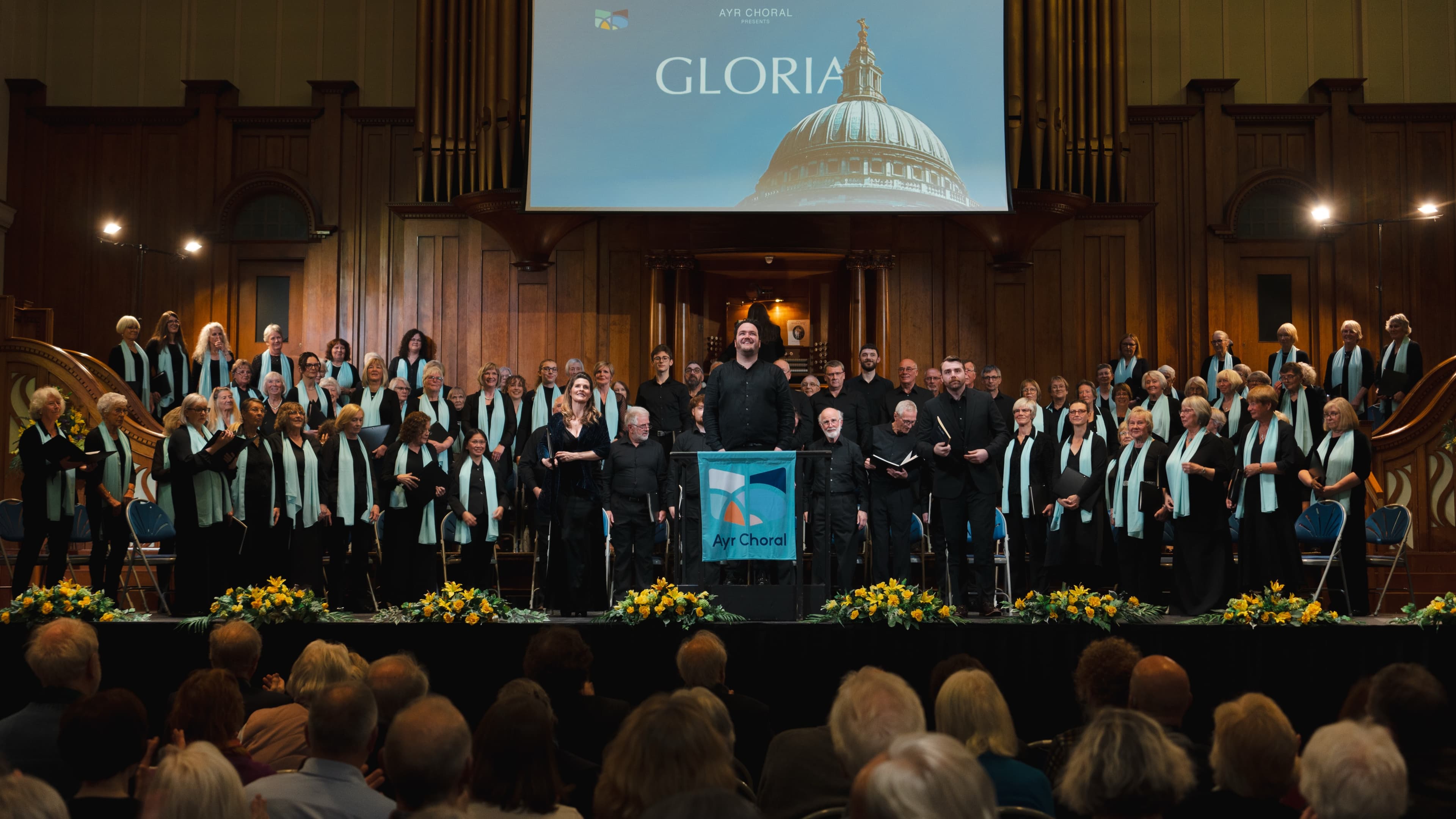 A full choir and soloists on stage beneath a title screen