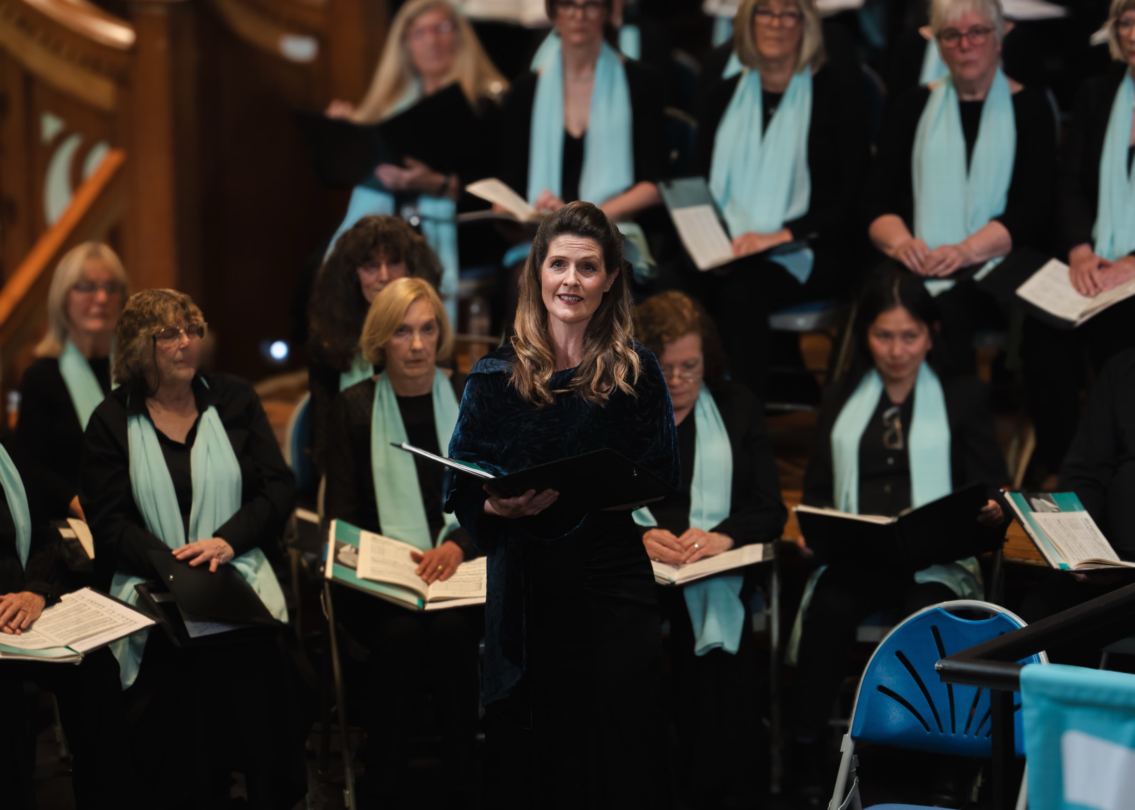 A soloist singing in front of a seated choir on a wood-panelled concert stage