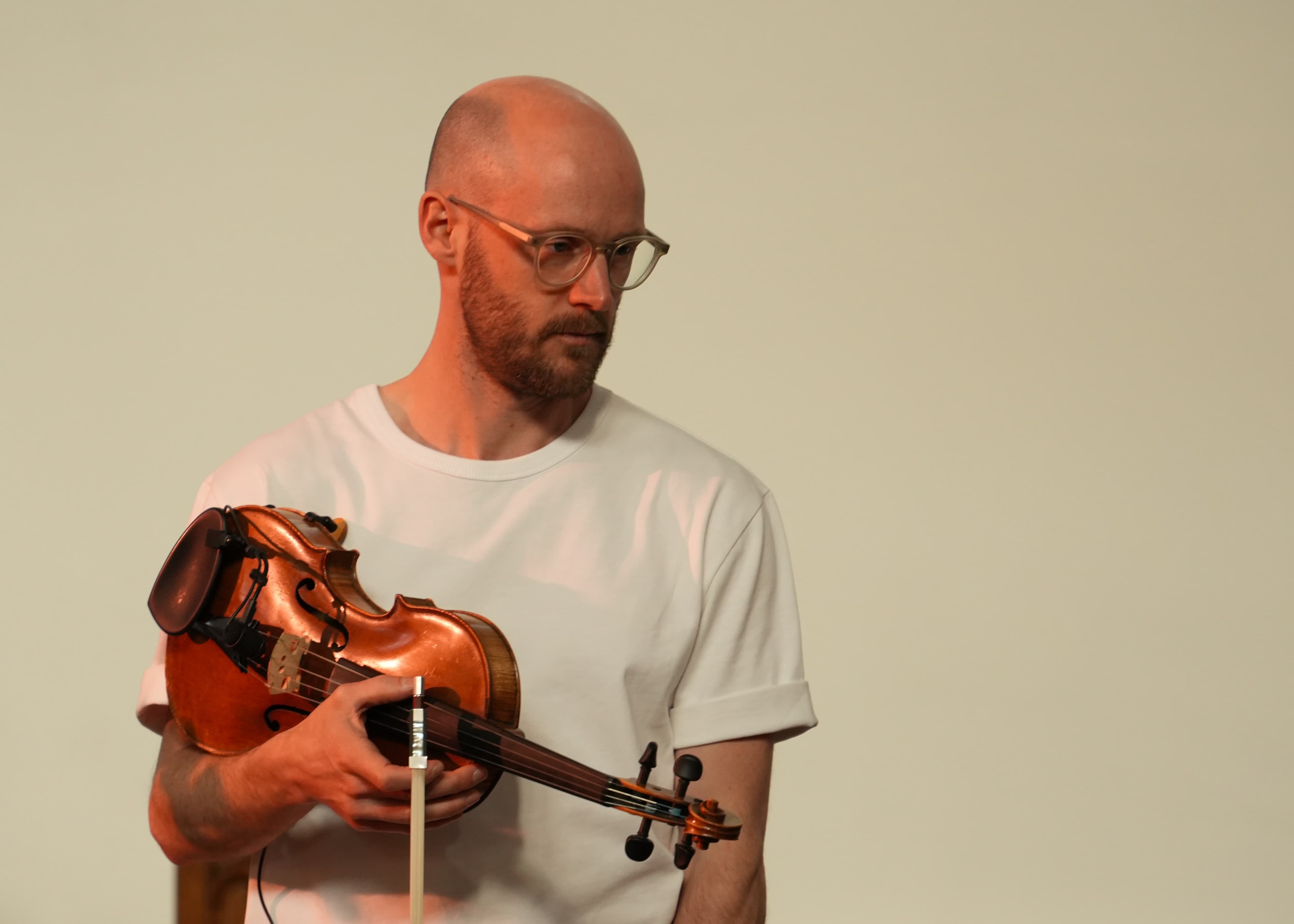 A portrait of a bald man in glasses holding a fretless violin against a plain cream background