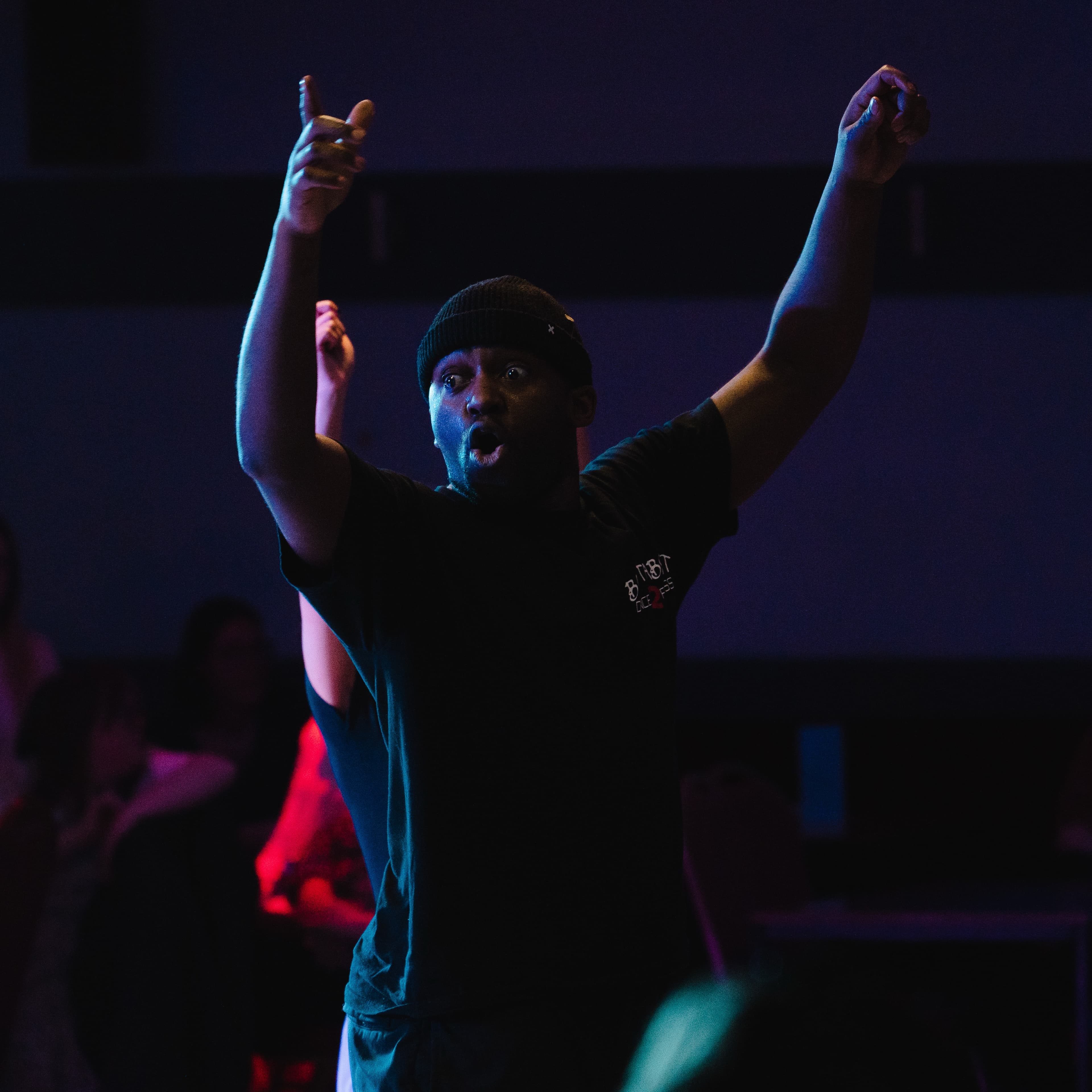 A performer in a beanie raising both arms with mouth open under blue stage lighting