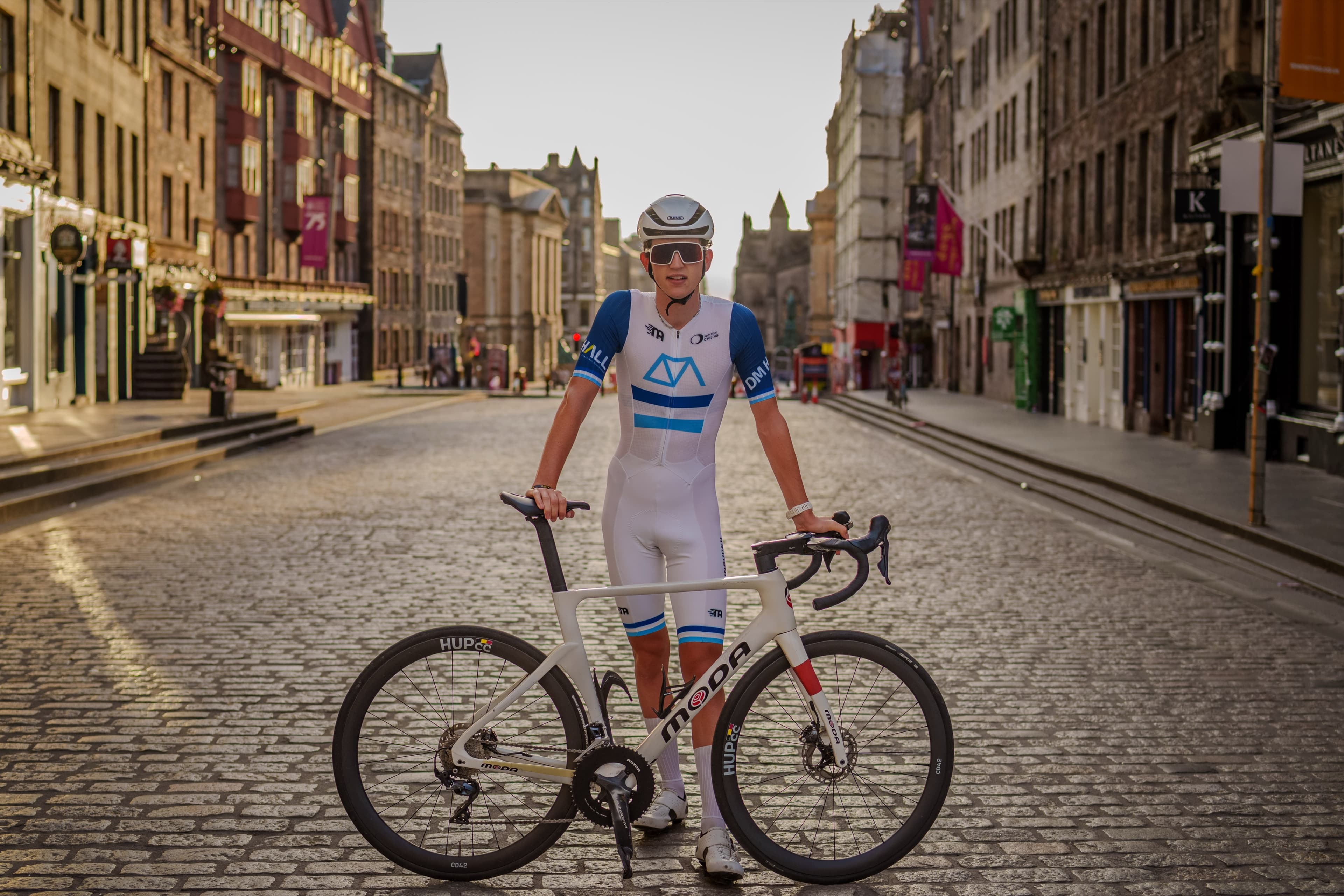 A cyclist in an aero skinsuit and helmet standing with his bike on a cobbled Edinburgh street at golden hour