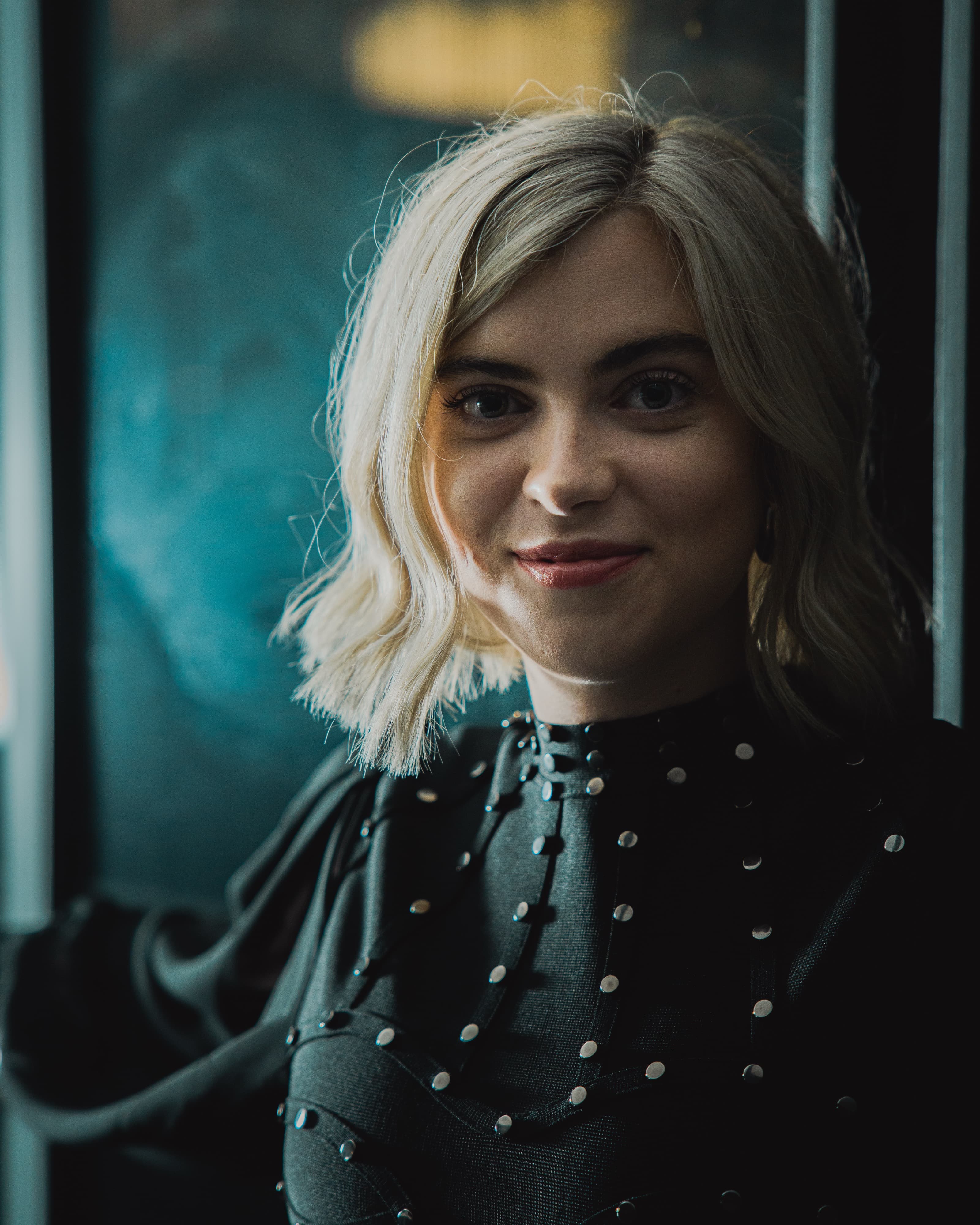 A portrait of a woman with blonde hair and a black studded top smiling softly against a dark teal background