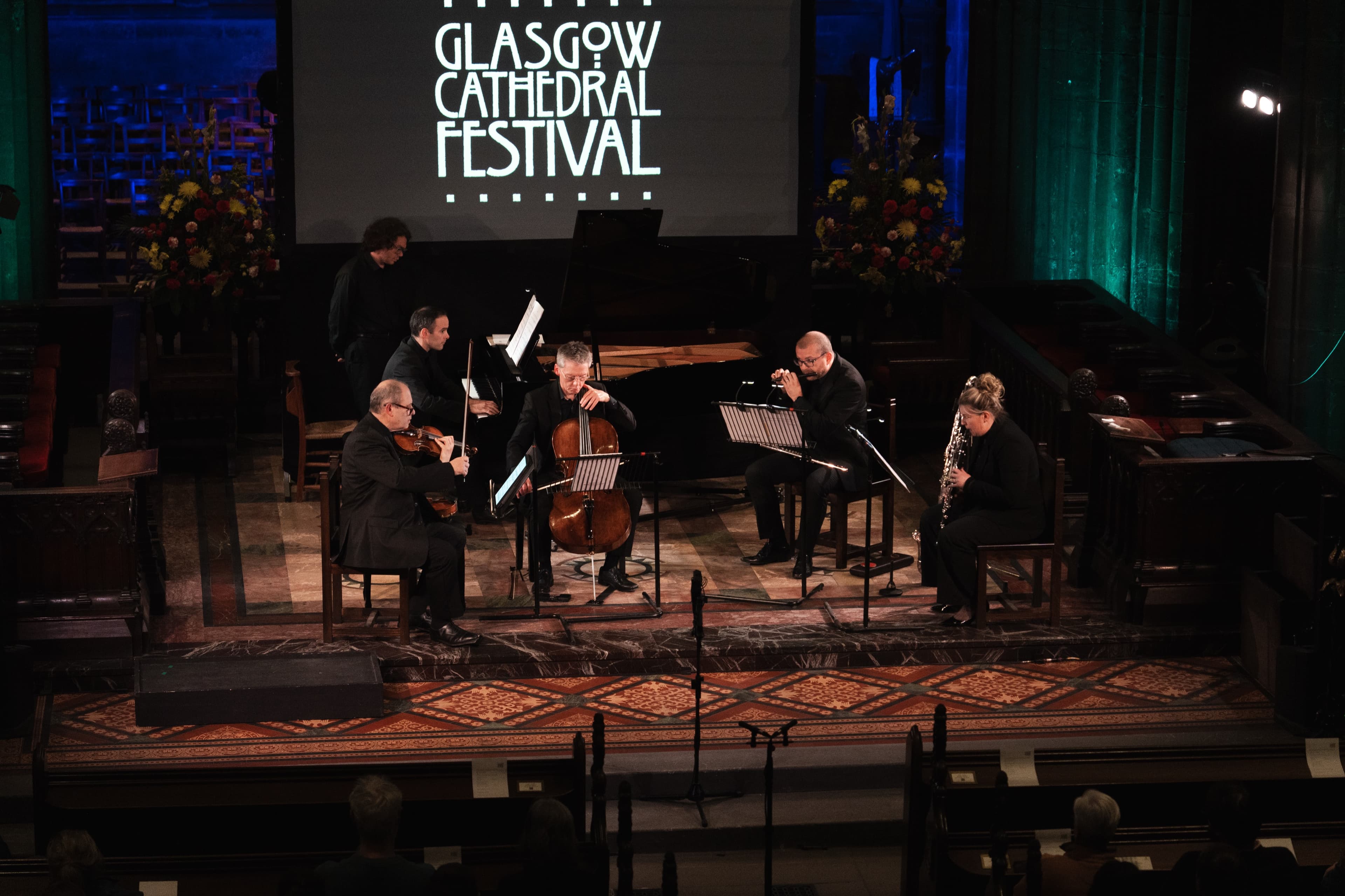A chamber ensemble of violin, cello, woodwind and piano performing together on a cathedral stage flanked by flower arrangements