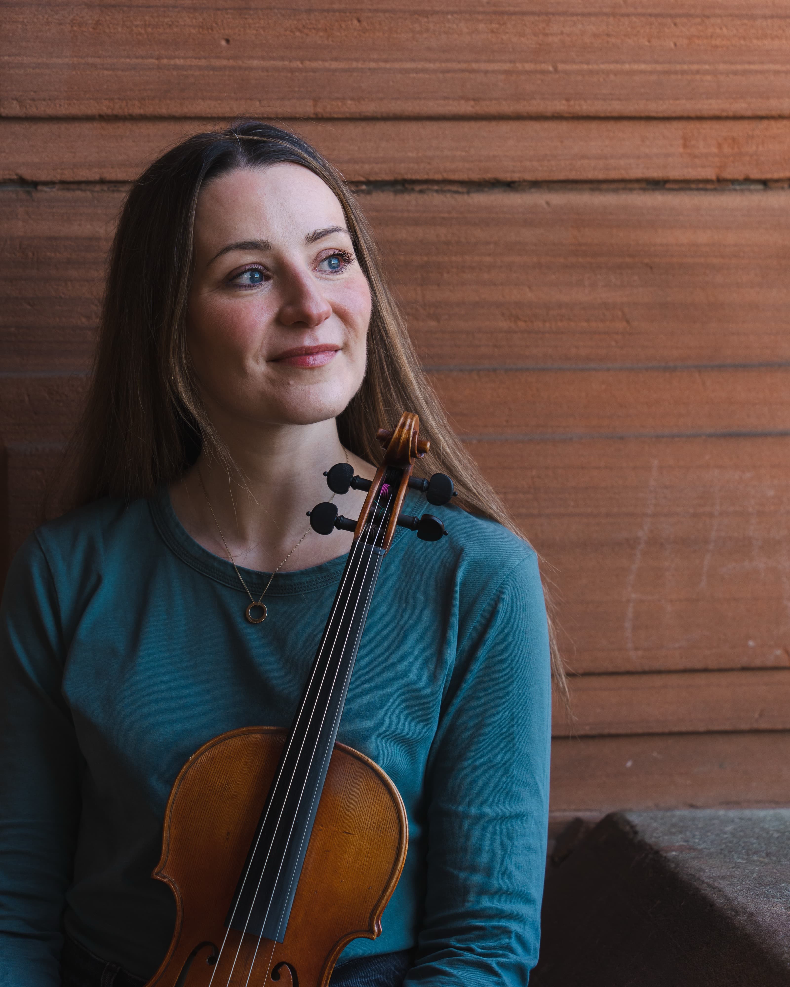 A smiling portrait of a violinist holding her instrument against a wooden wall