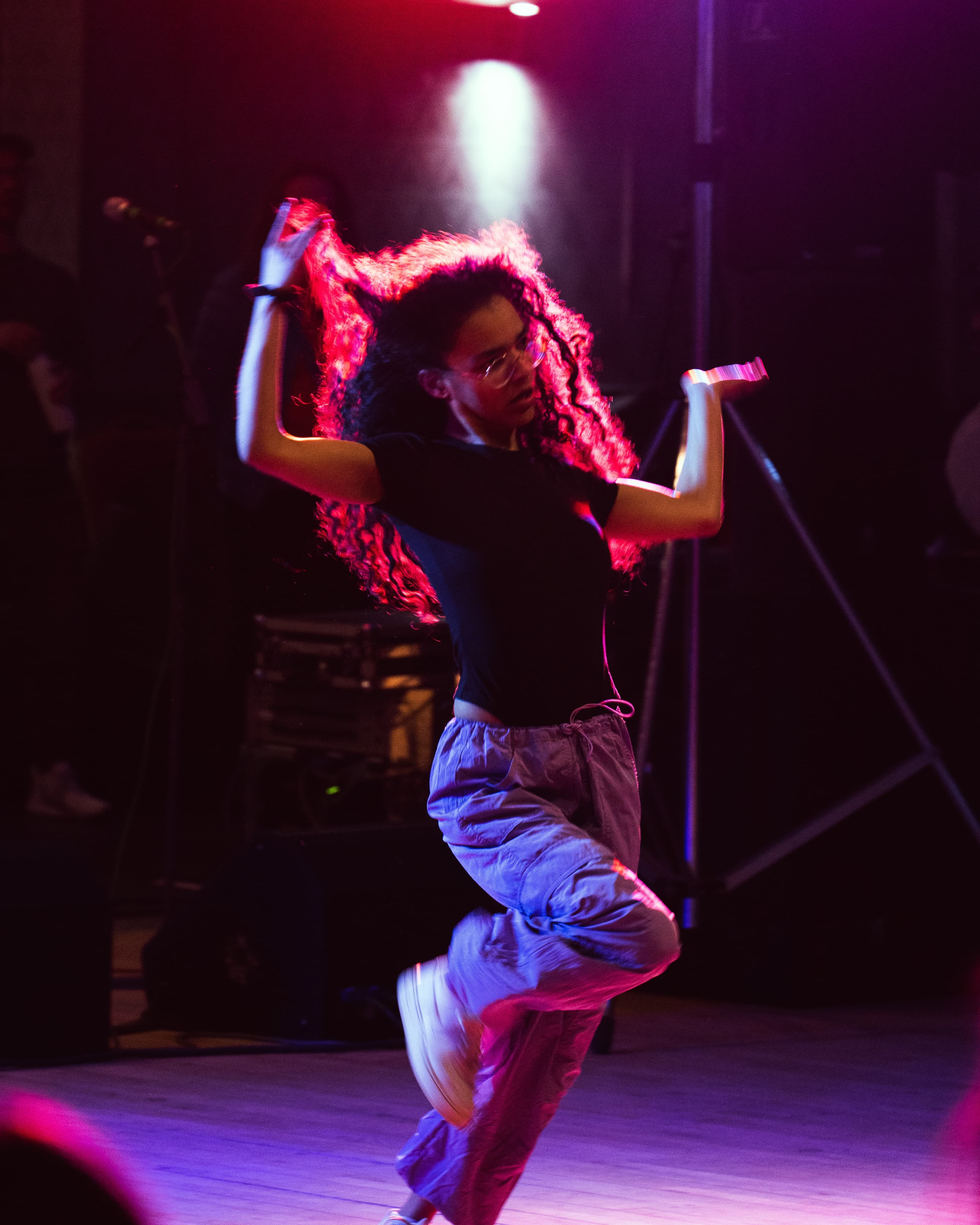 A dancer with wind-blown curly hair captured mid-jump under pink stage lighting