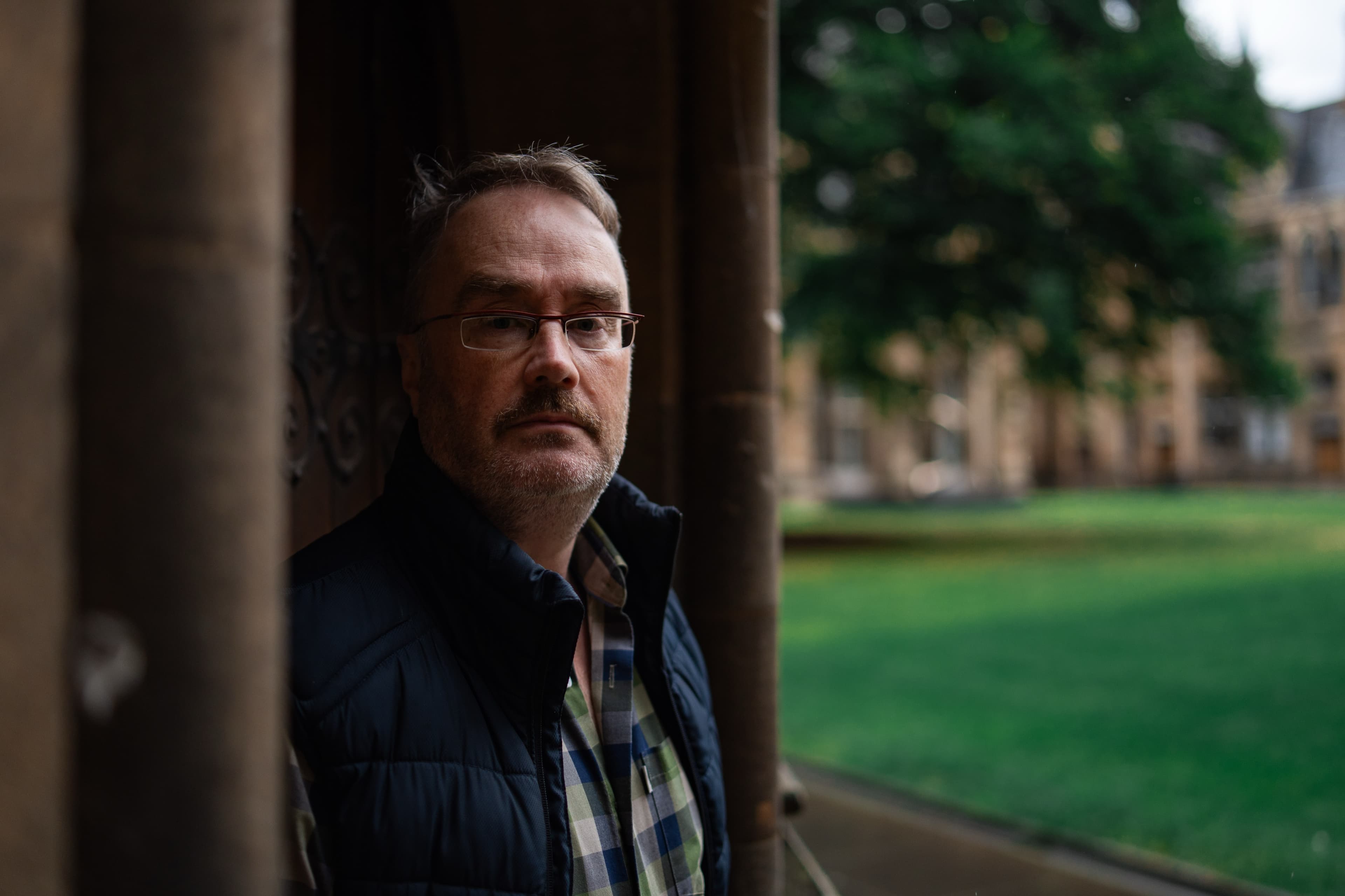A portrait of a man in glasses and a gilet standing in a stone archway overlooking a lawn