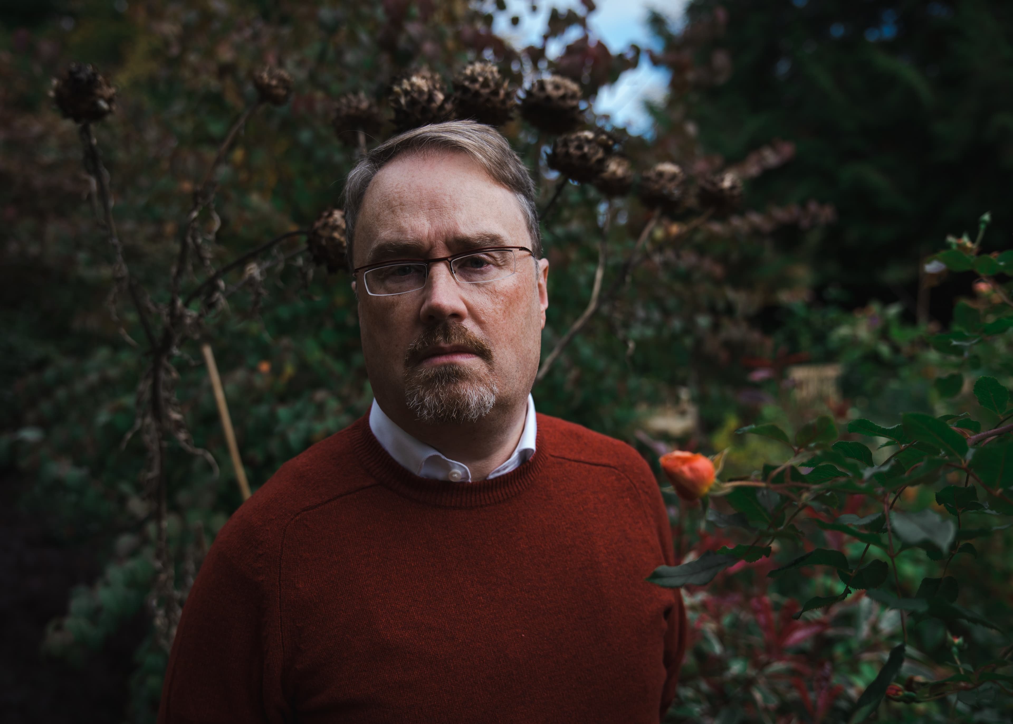 A portrait of a man in a rust-coloured jumper standing in a garden