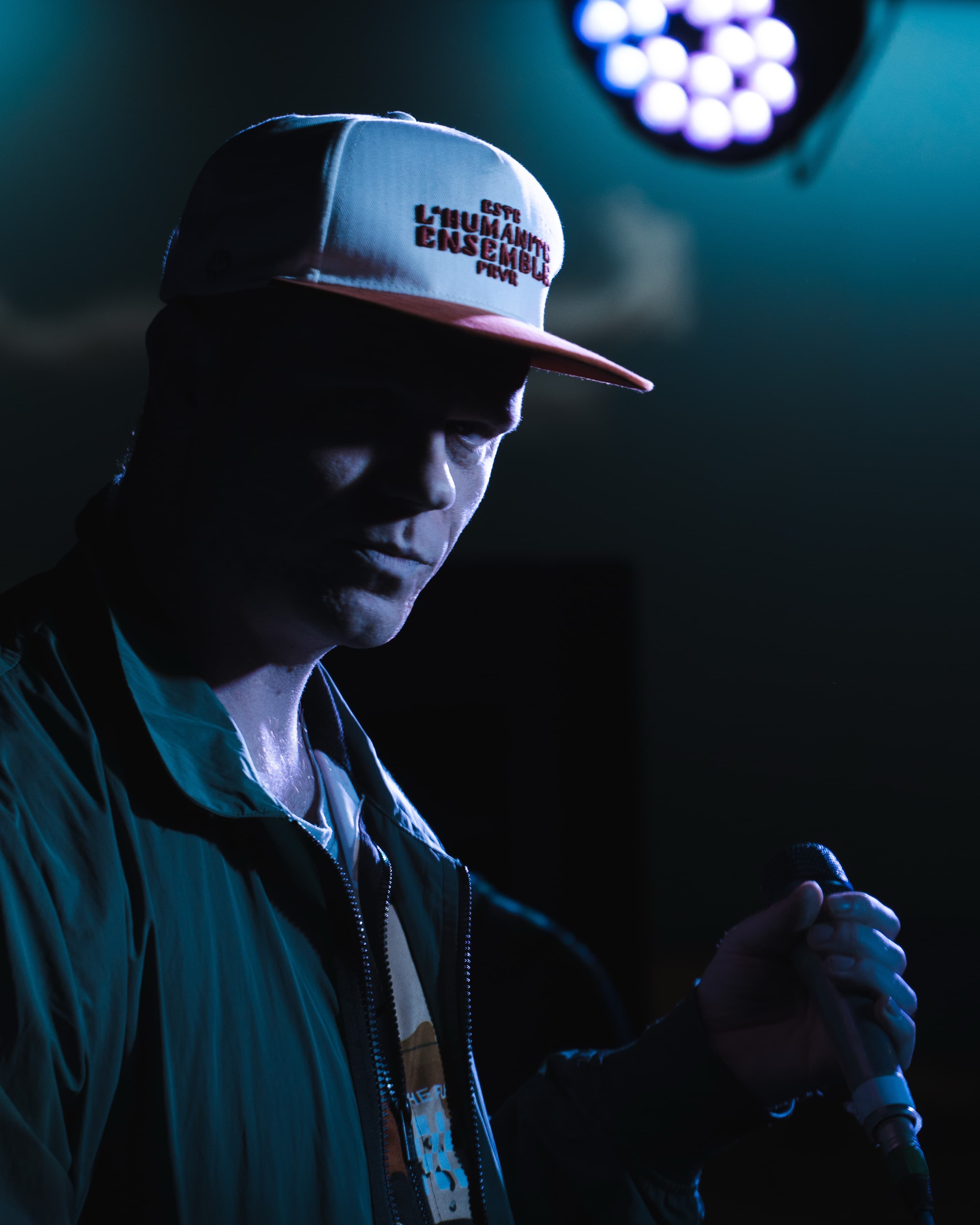 A moody portrait of a man in a cap holding a microphone under teal stage lighting