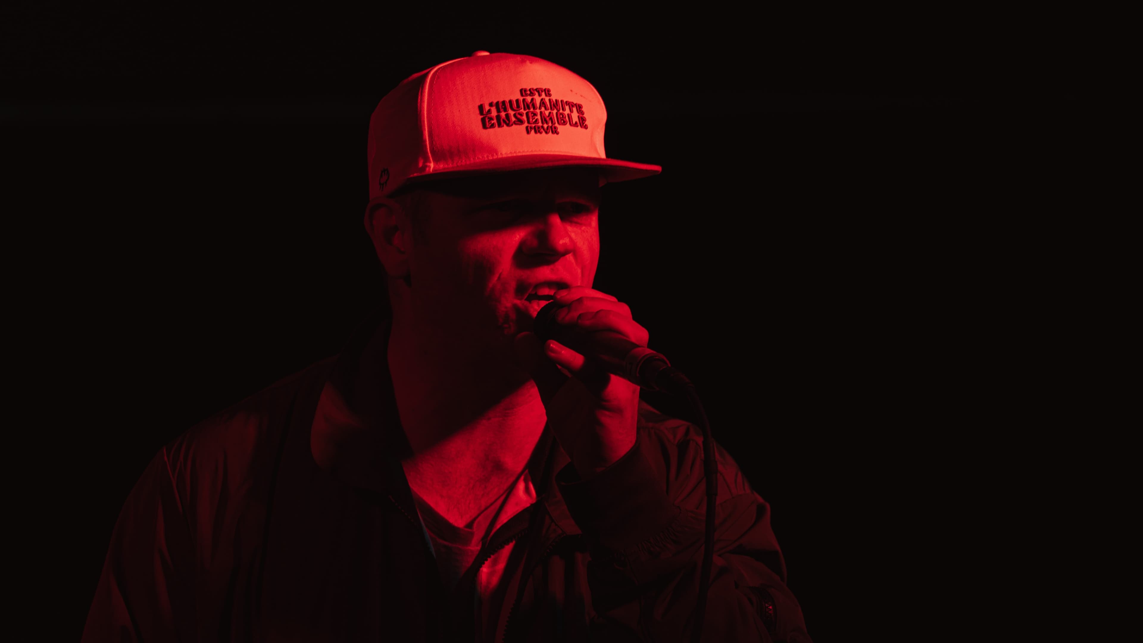 A performer in a printed cap holding a microphone under red stage lighting