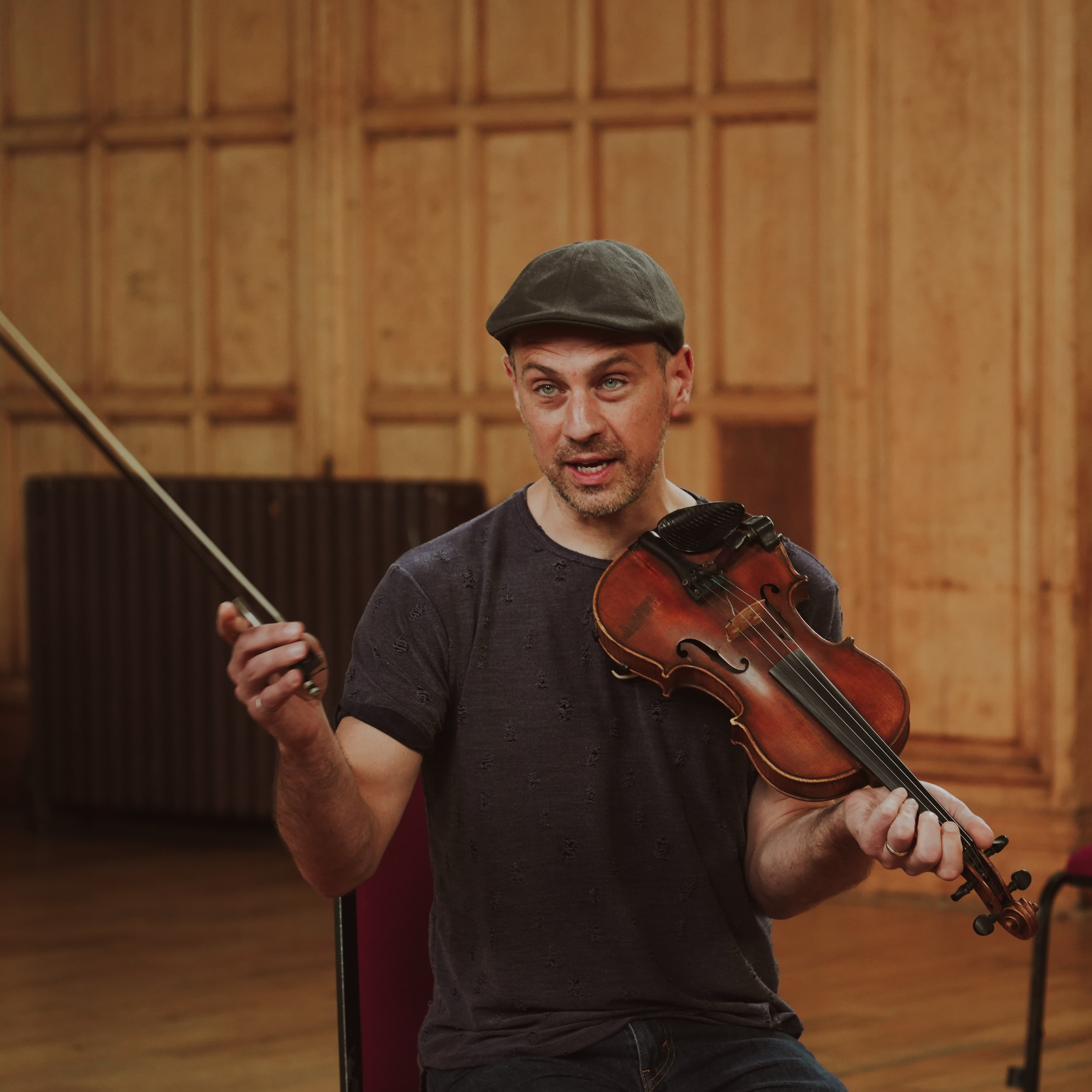 A fiddle player in a flat cap speaking to onlookers while holding his violin and bow