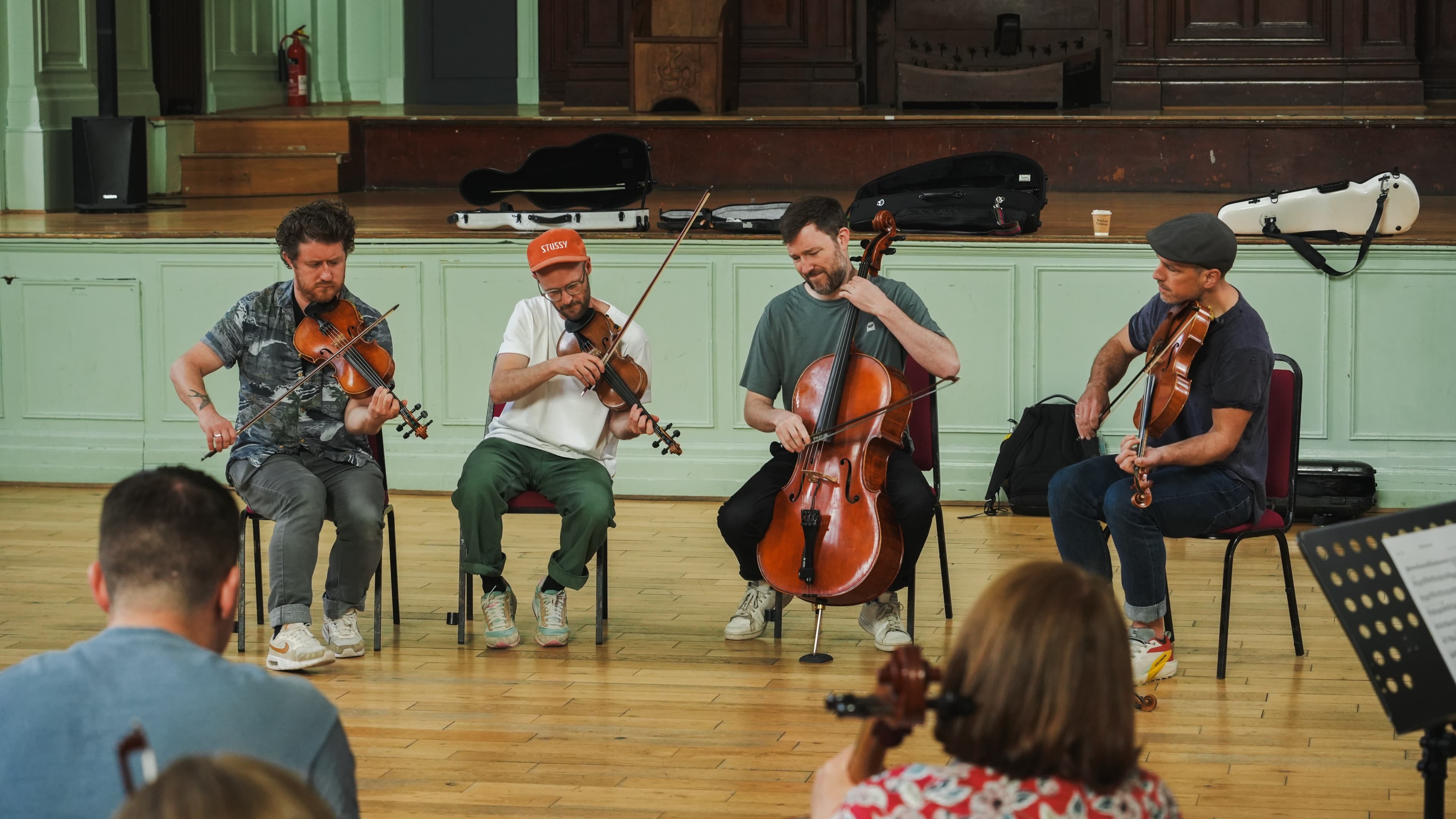 Four musicians playing violins and cello together in a row inside a wood-paneled hall