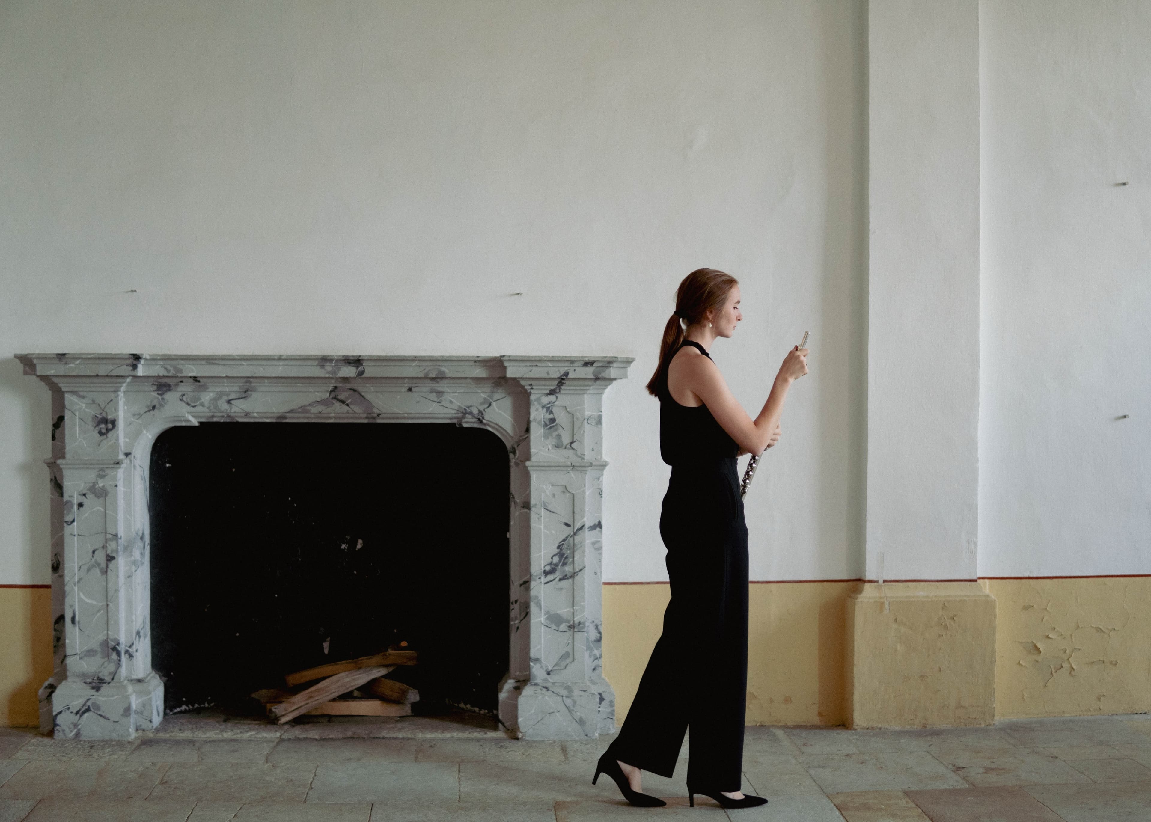 A flautist standing in profile beside an ornate marble fireplace, holding her flute