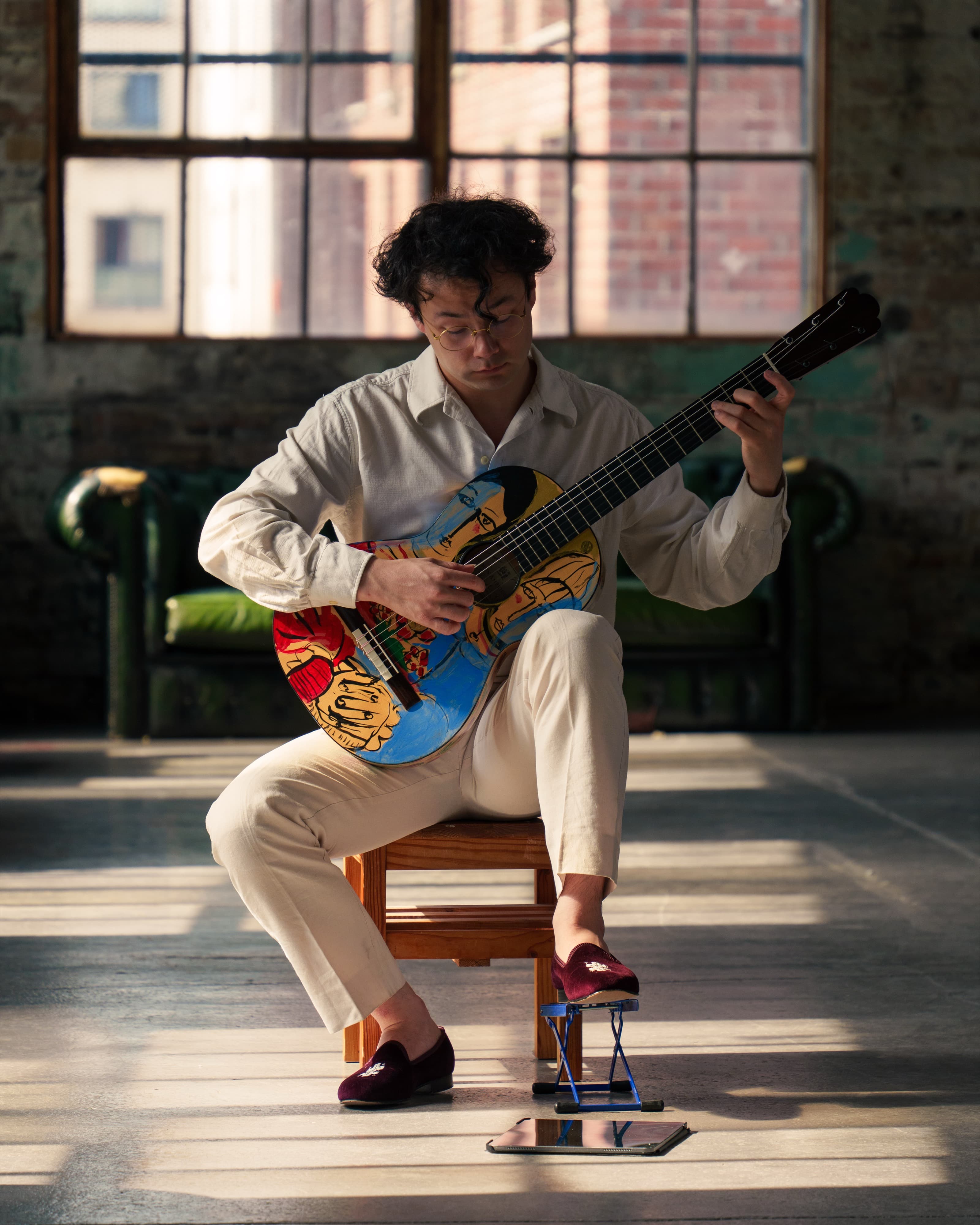 A guitarist playing a hand-painted classical guitar seated in a sunlit industrial loft