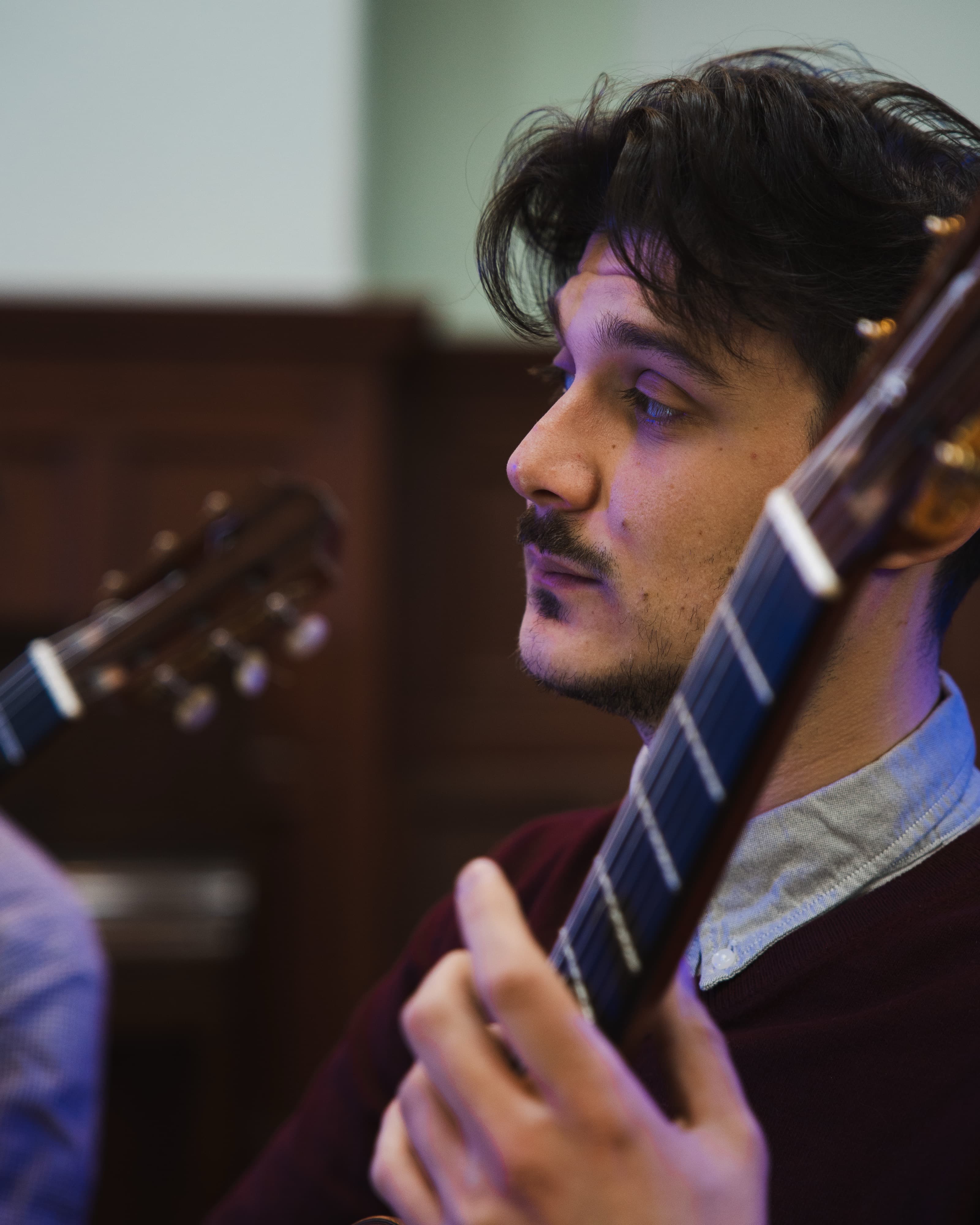 A close-up profile portrait of a man playing classical guitar, guitar neck in the foreground, moody purple stage lighting