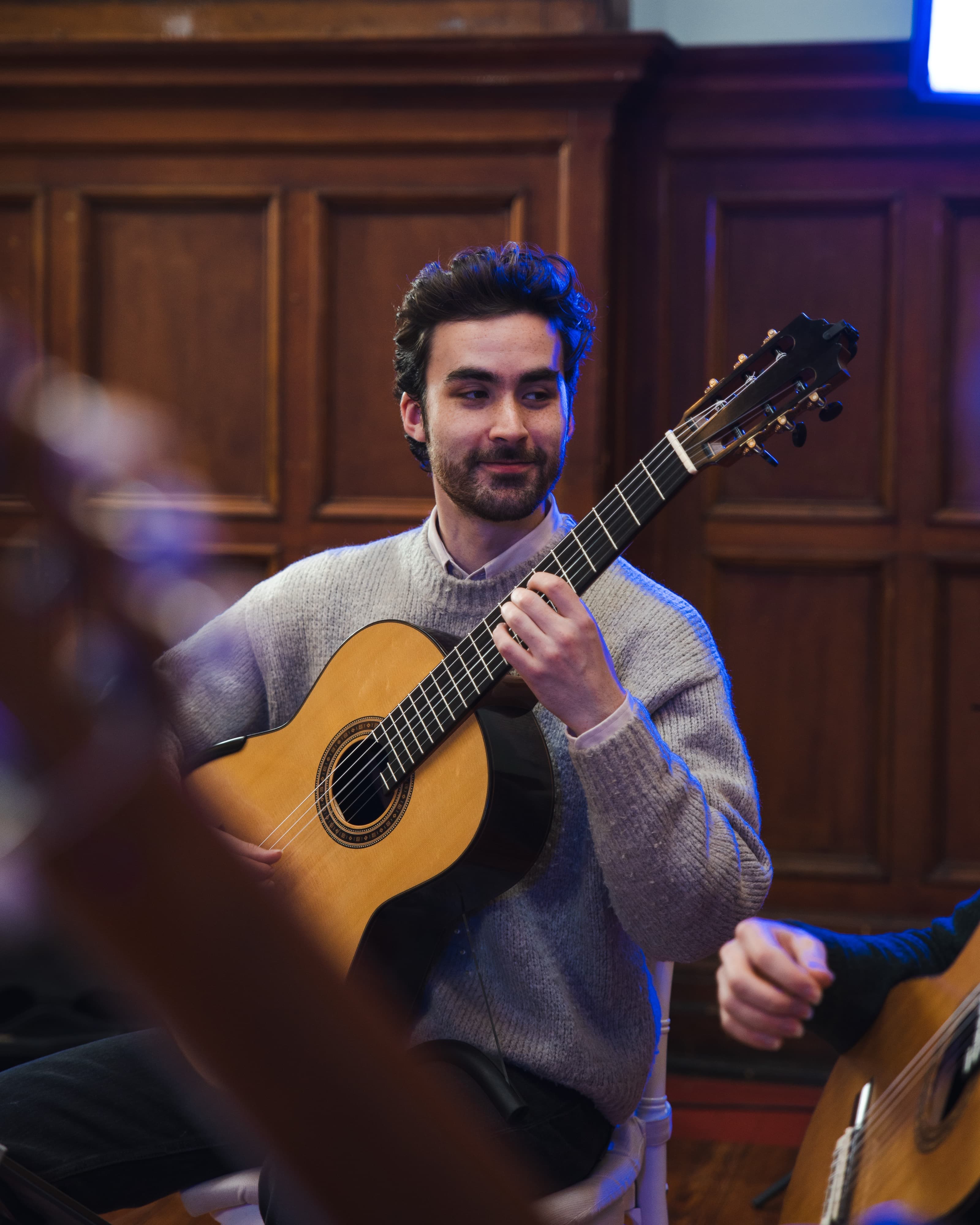 A portrait of a man smiling while playing classical guitar in a wood-panelled room