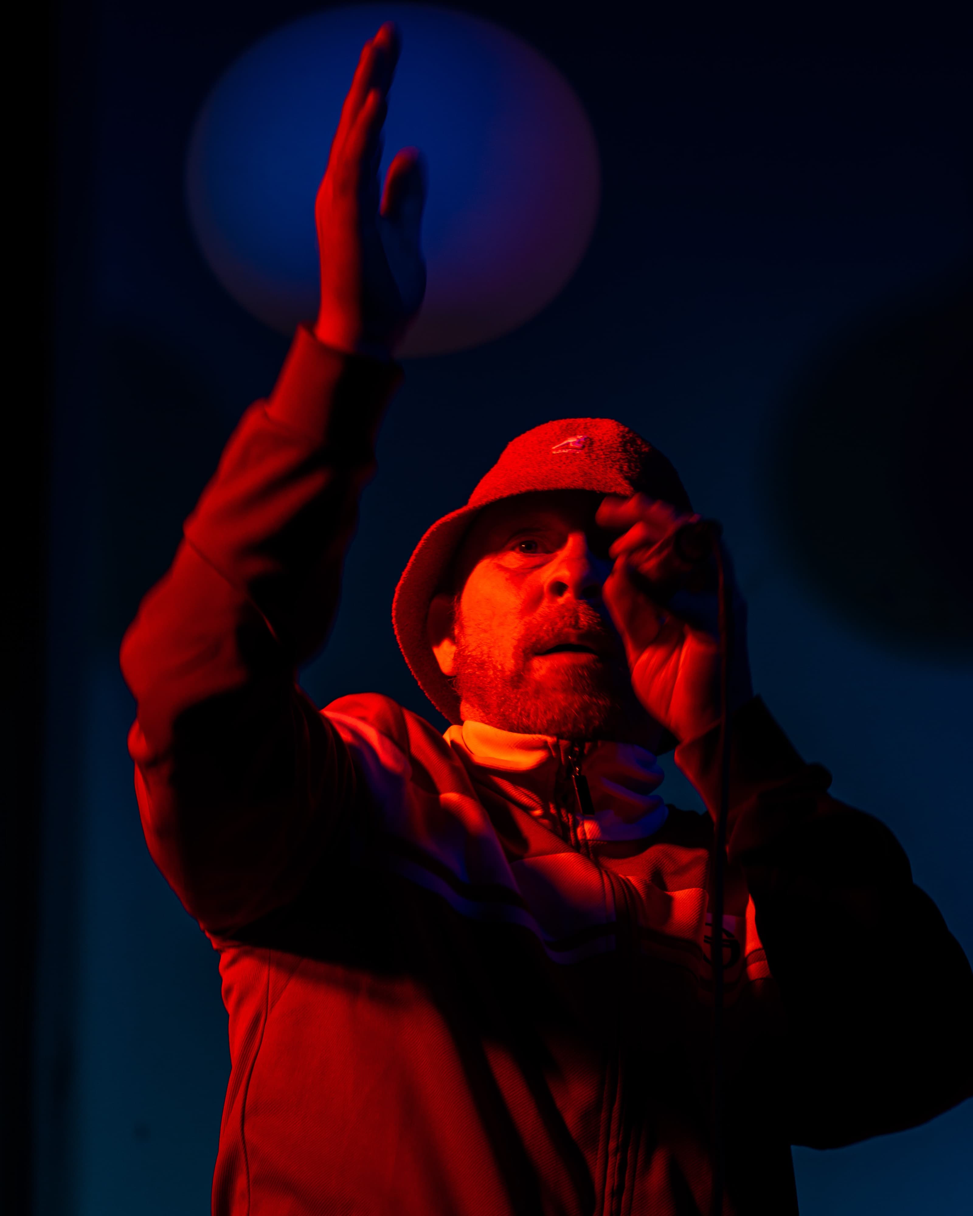 A performer in a red bucket hat raising a hand while holding a microphone, under red and blue stage lighting