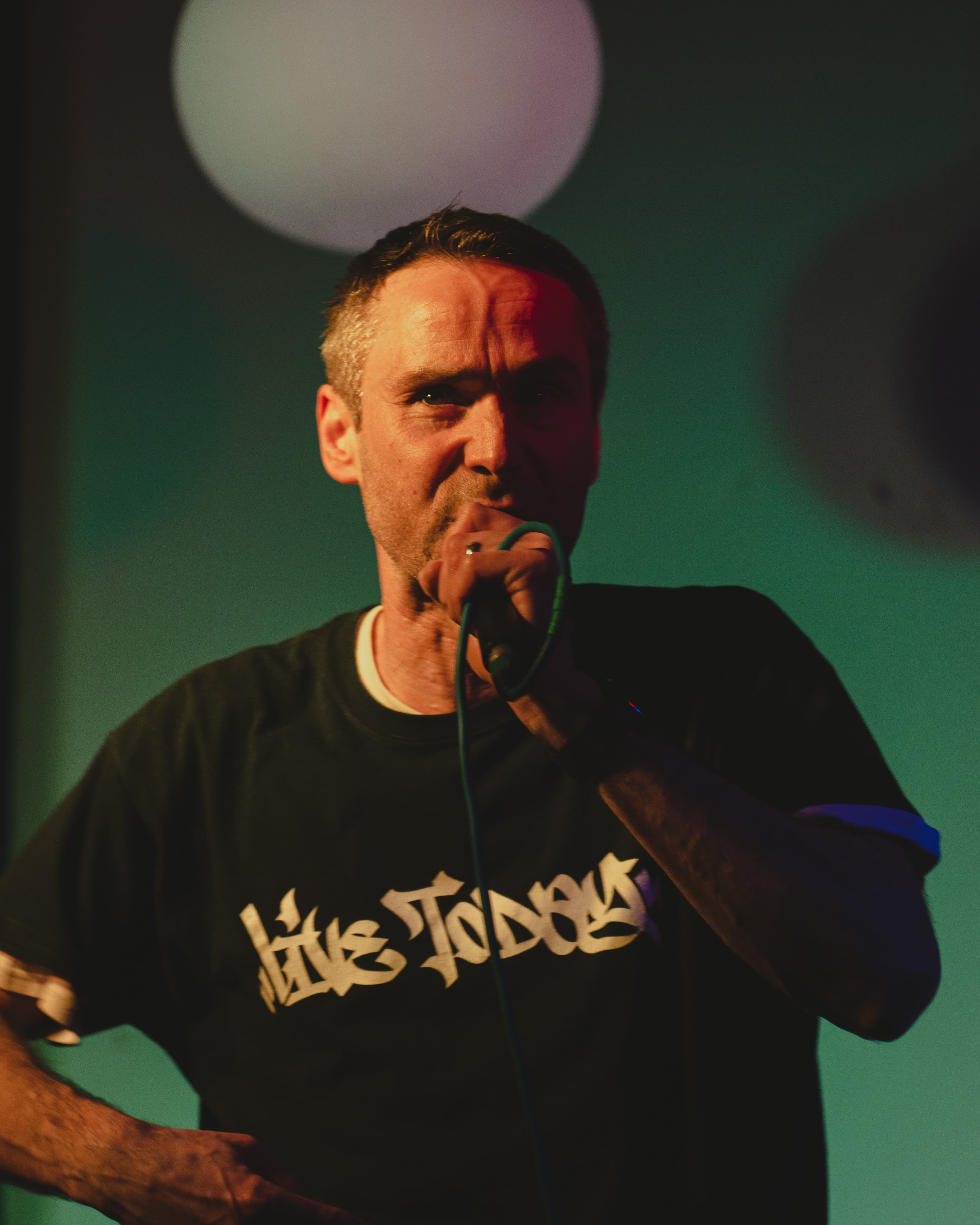 A performer in a red bucket hat singing into a microphone under red and blue stage lighting