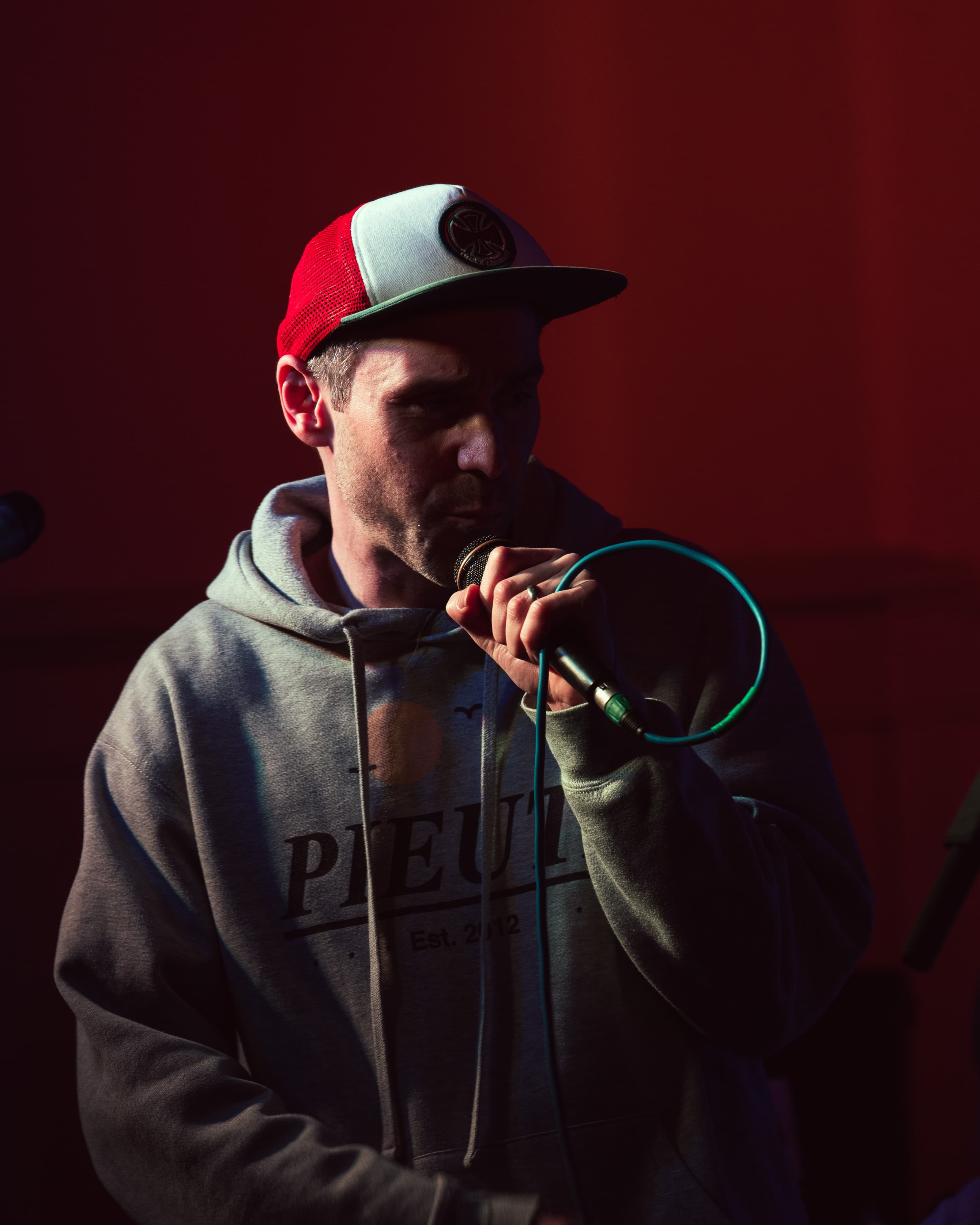 A performer in a trucker cap and hoodie holding a microphone against a red backdrop
