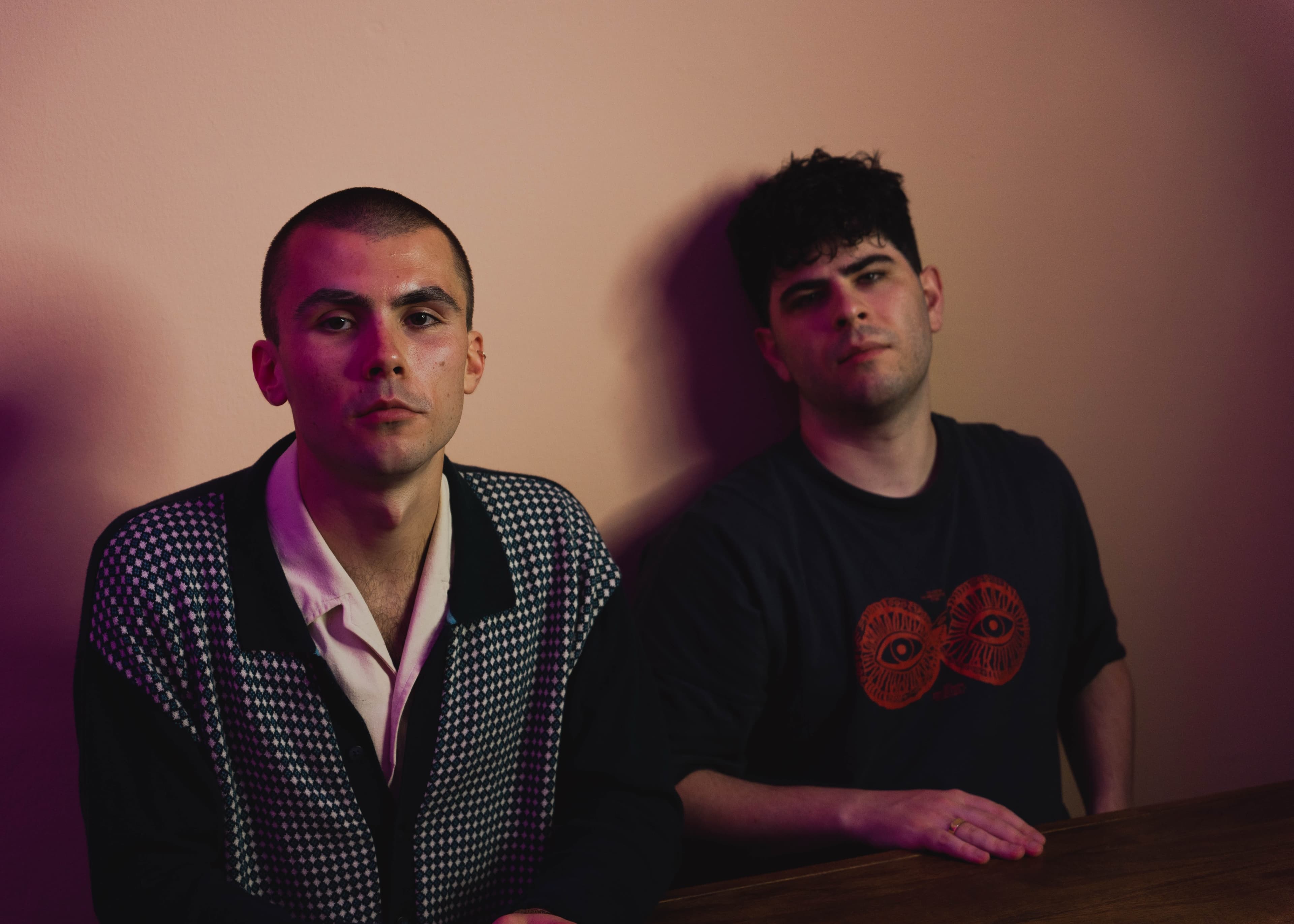 Two men seated together under pink lighting, one in a patterned jacket, both looking at camera