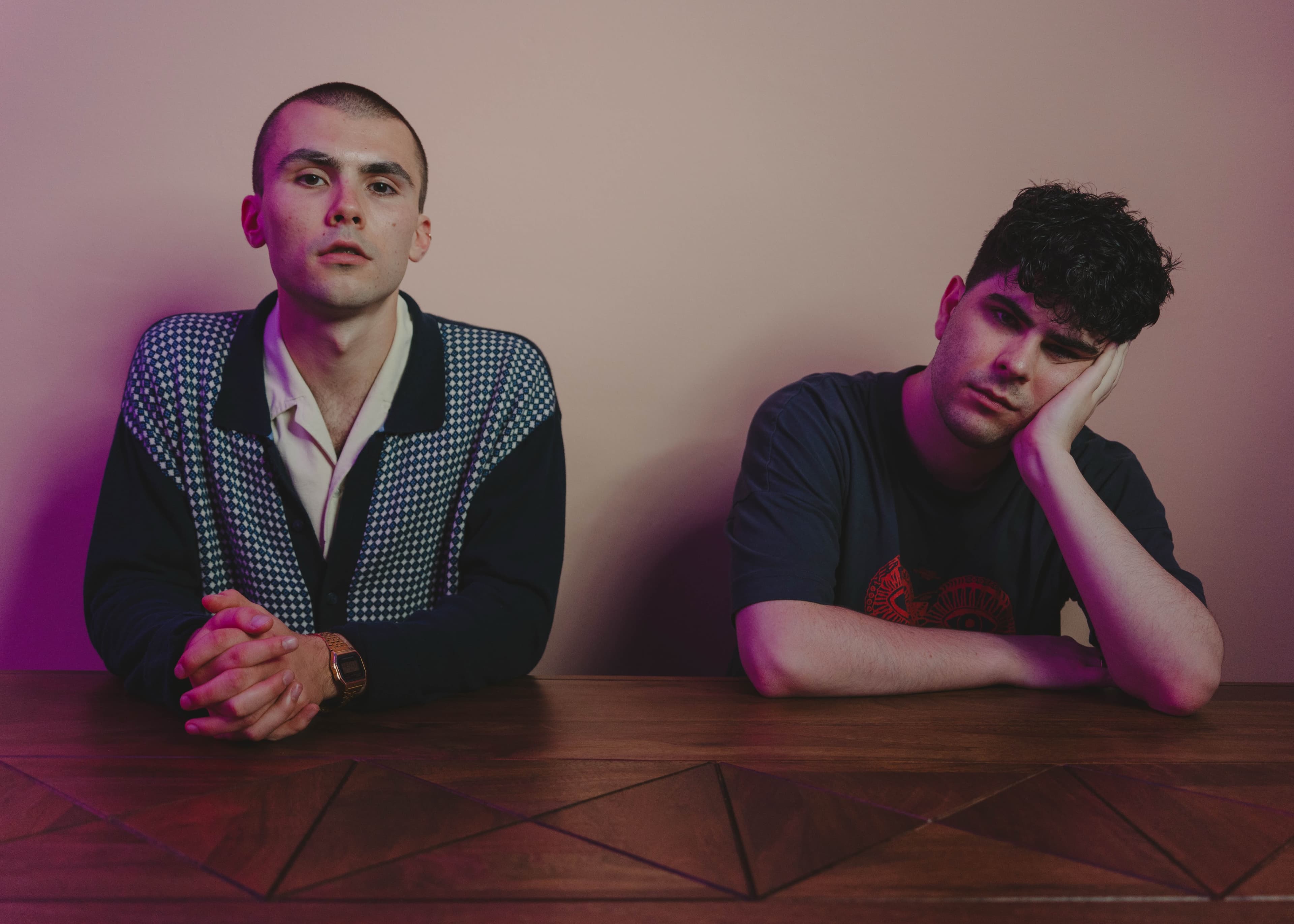 Two men leaning on a table under pink and purple lighting, one resting his head on his hand