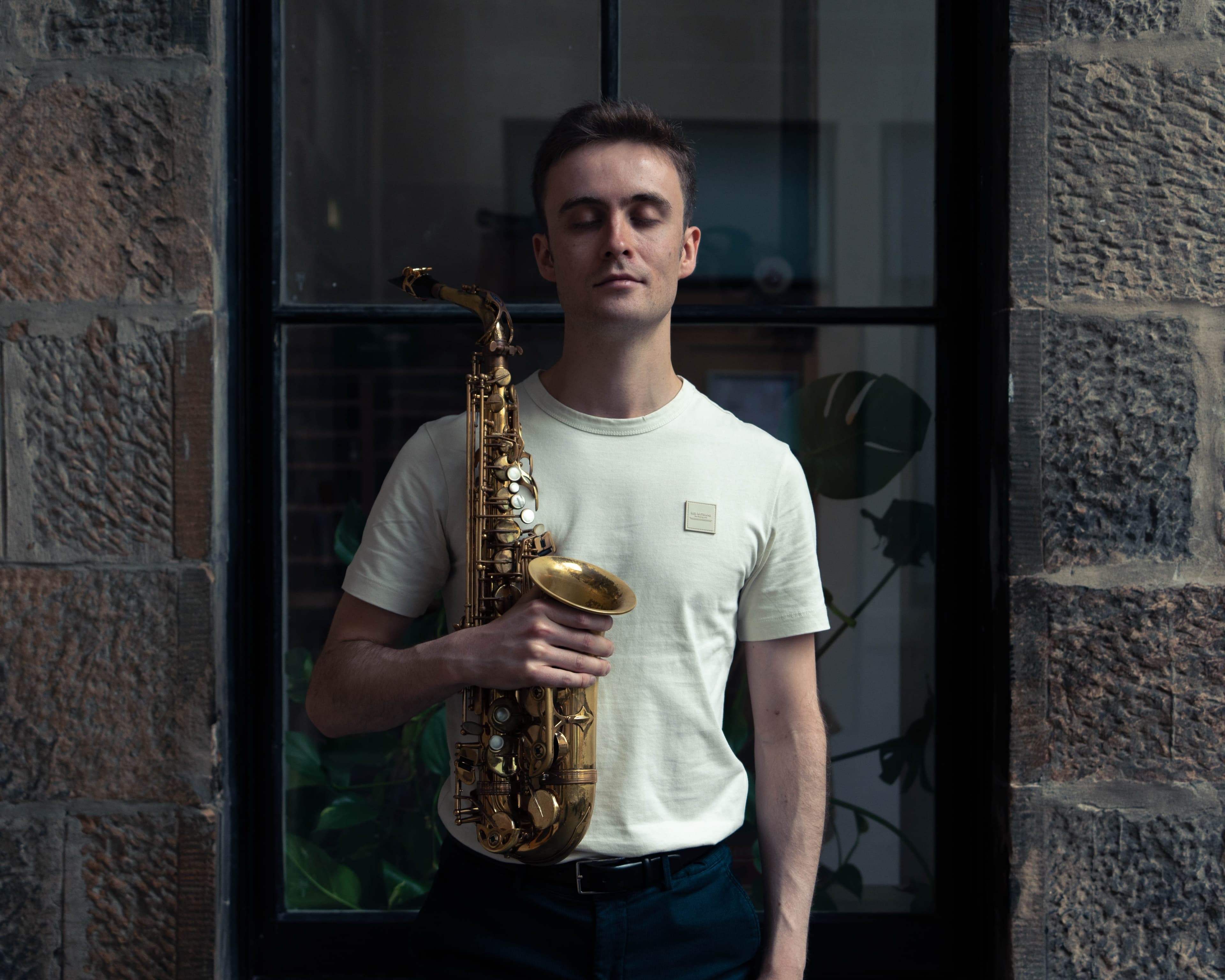 A portrait of a musician with eyes closed holding a saxophone against a stone wall