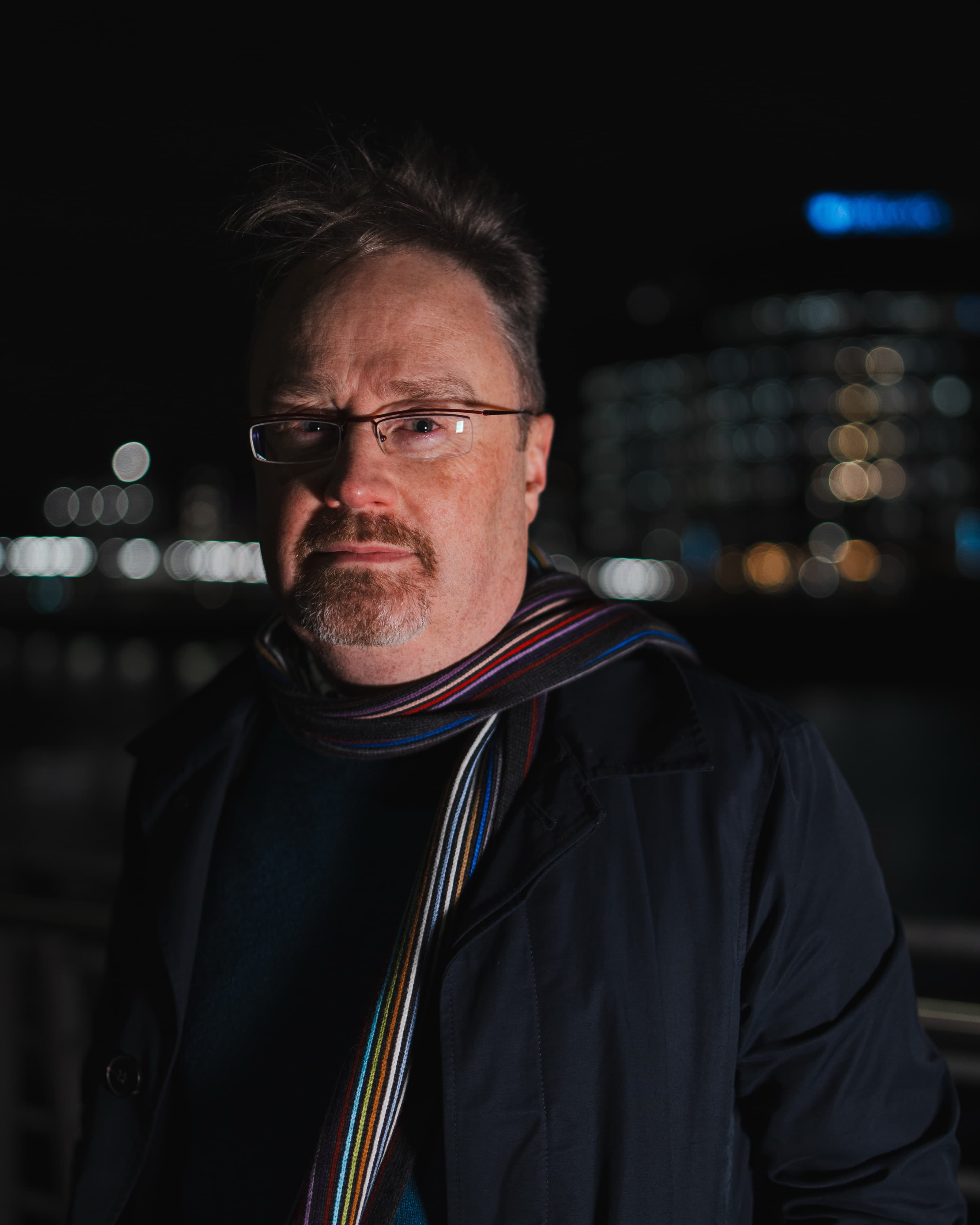 A portrait of a man in glasses and a striped scarf against a blurred night-time city skyline