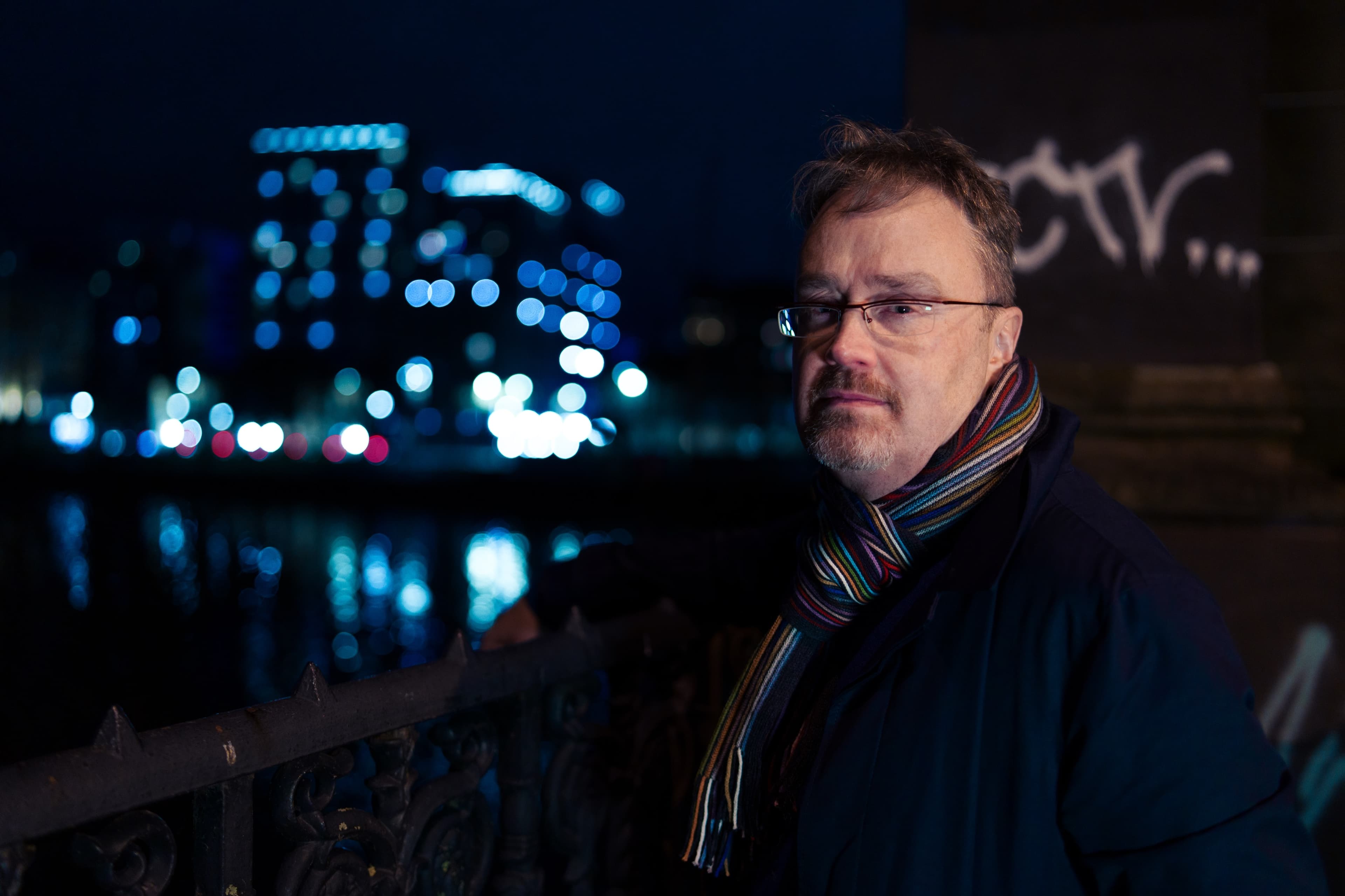 A man in glasses and a striped scarf looking off to the side beside a railing at night, city lights and graffiti in the background
