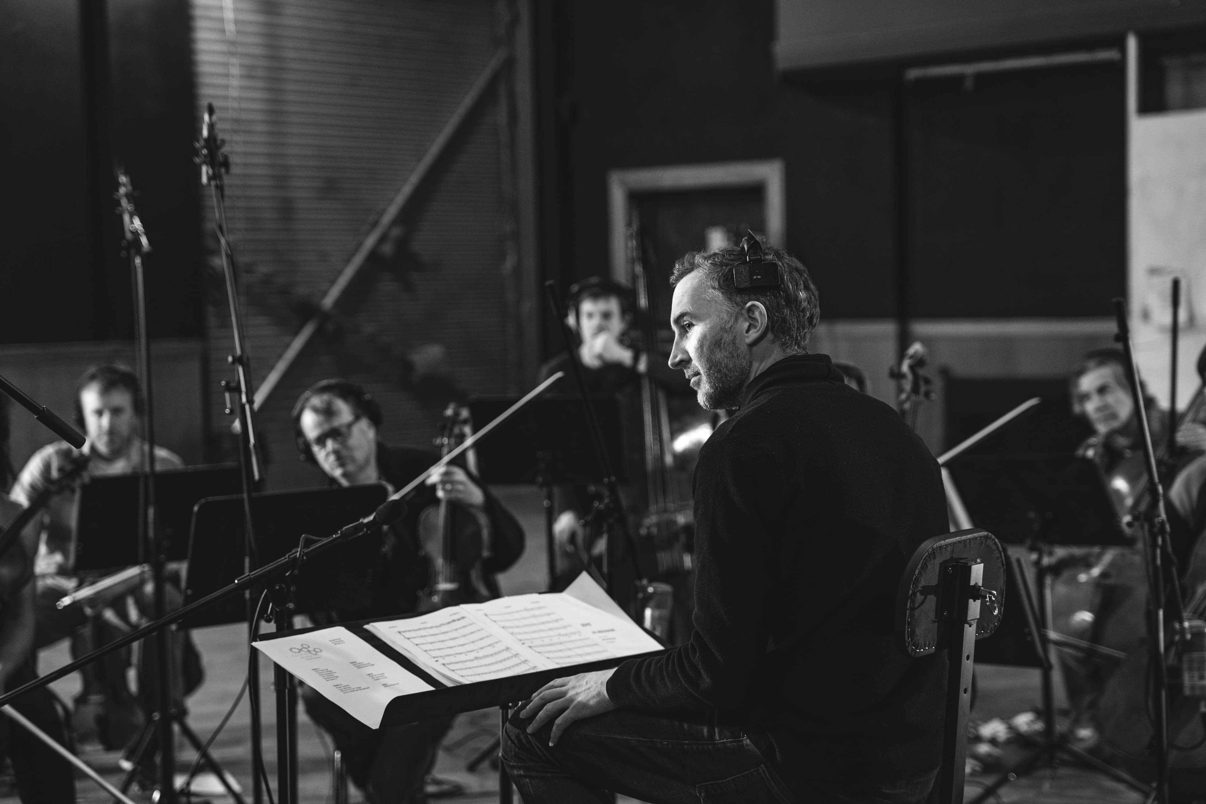 A man reading sheet music beside string musicians in a recording session, black and white