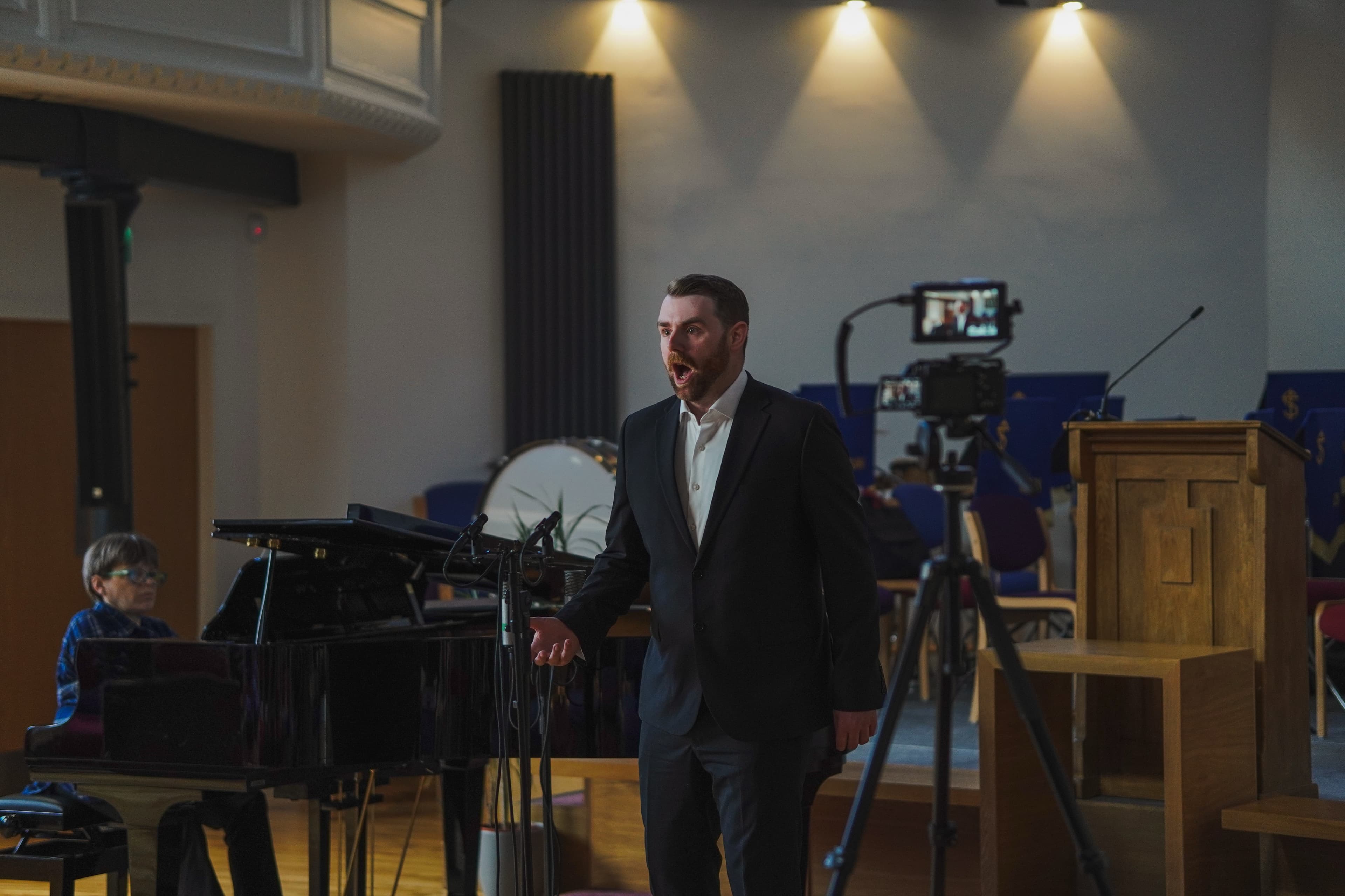 A singer in a suit performing beside a grand piano with a pianist and camera in the background