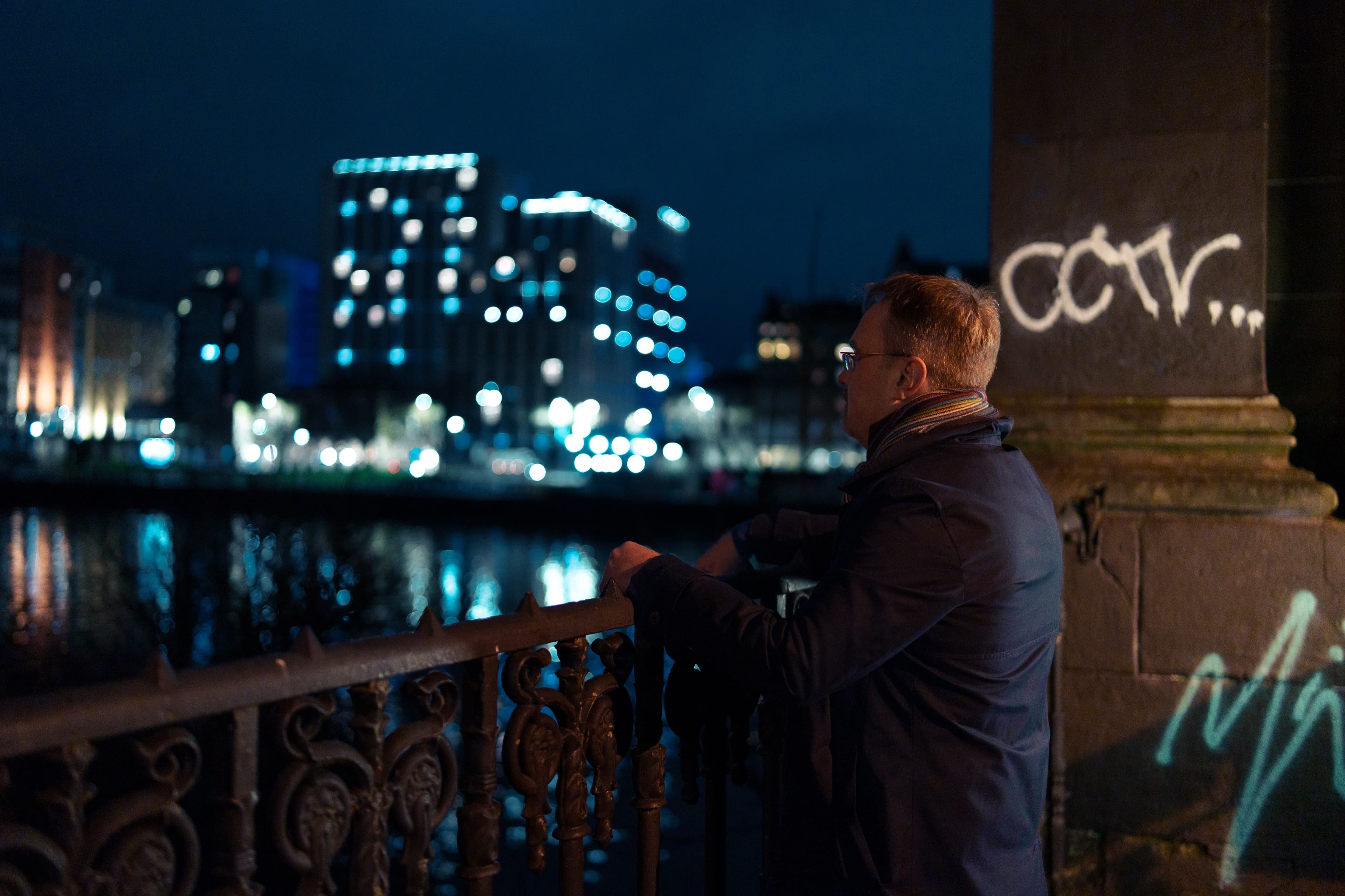 A man leaning on a railing beside a river at night looking towards an illuminated city skyline, with graffiti visible on a stone pillar beside him