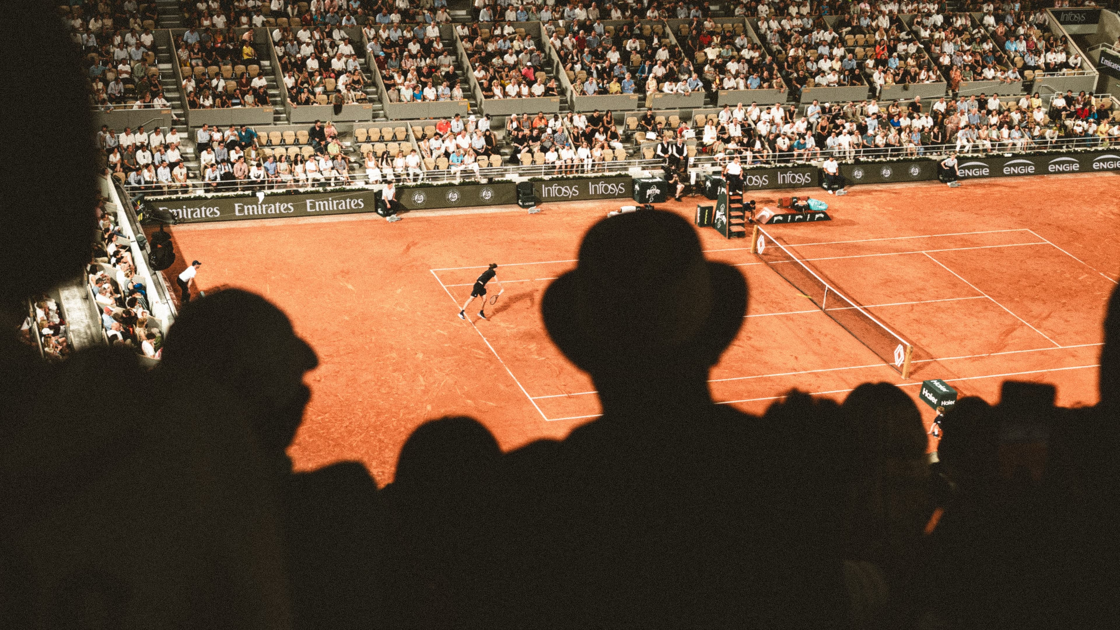 Silhouetted spectators watching a tennis match on a sunlit clay court at Roland Garros