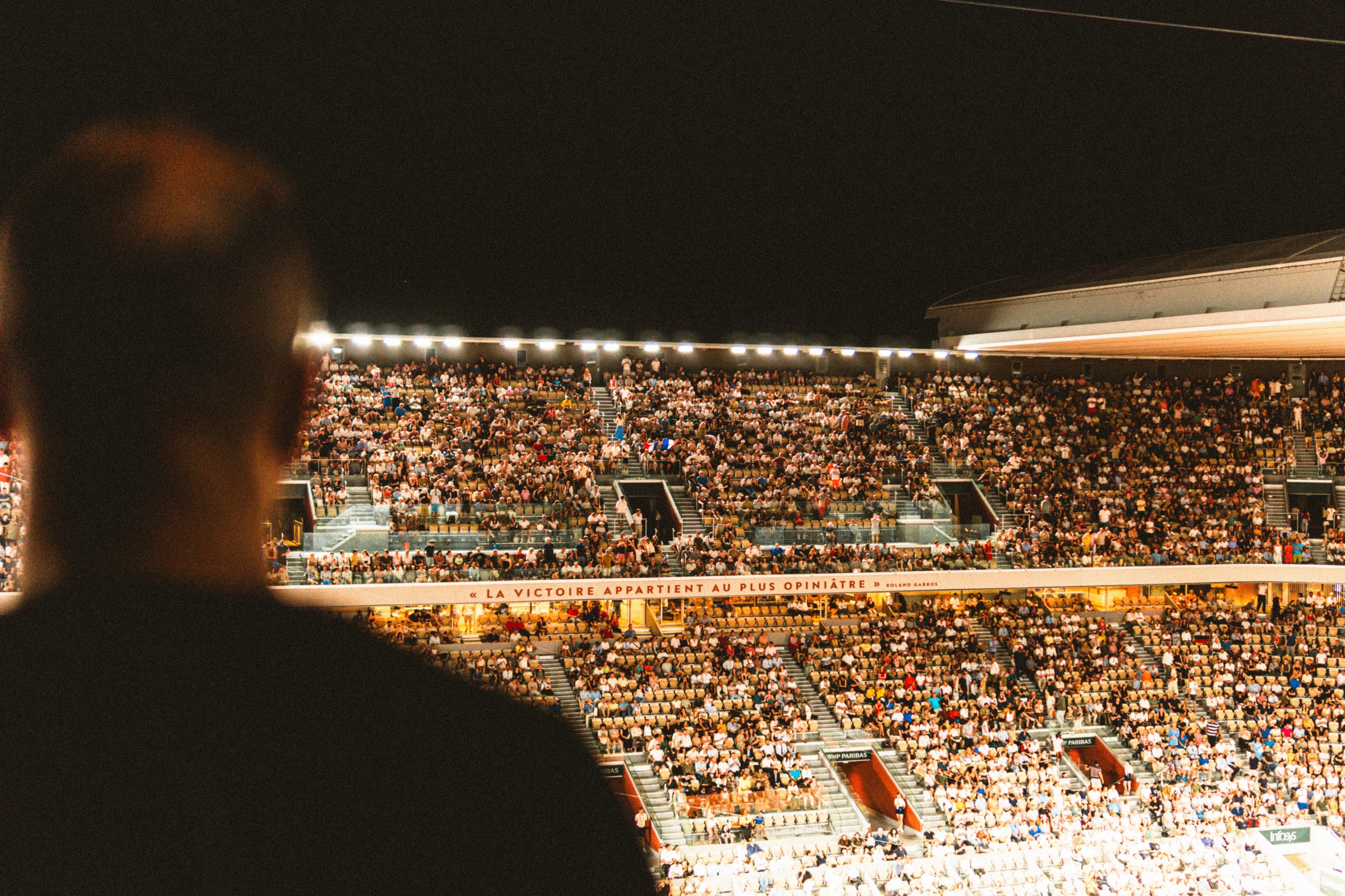 Silhouetted spectator overlooking a packed floodlit stadium crowd at night, Roland Garros