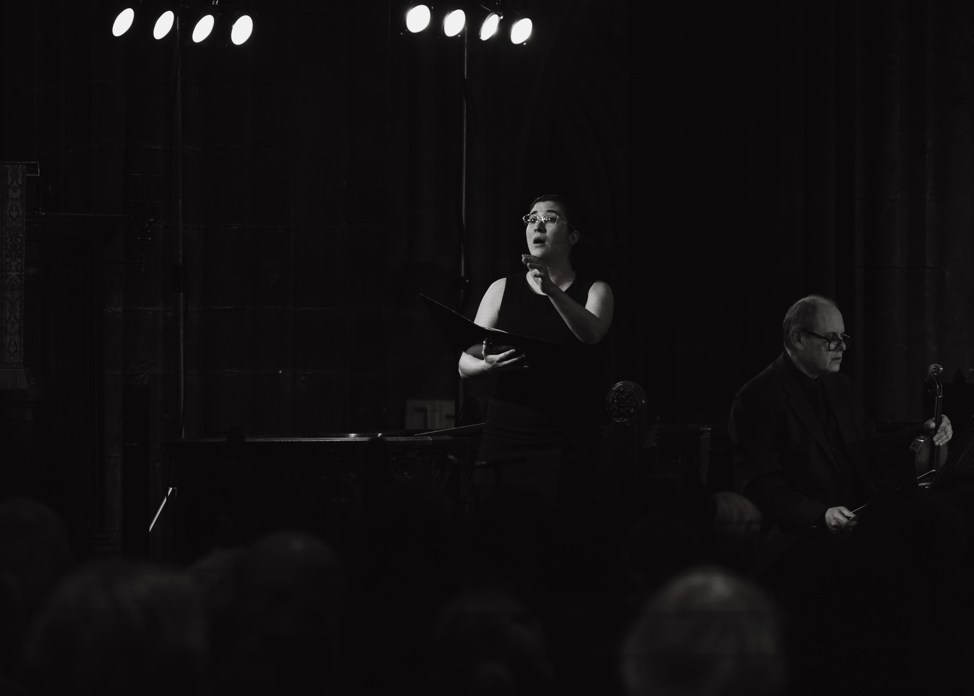 A singer holding sheet music and gesturing while performing, with a violinist nearby, black and white