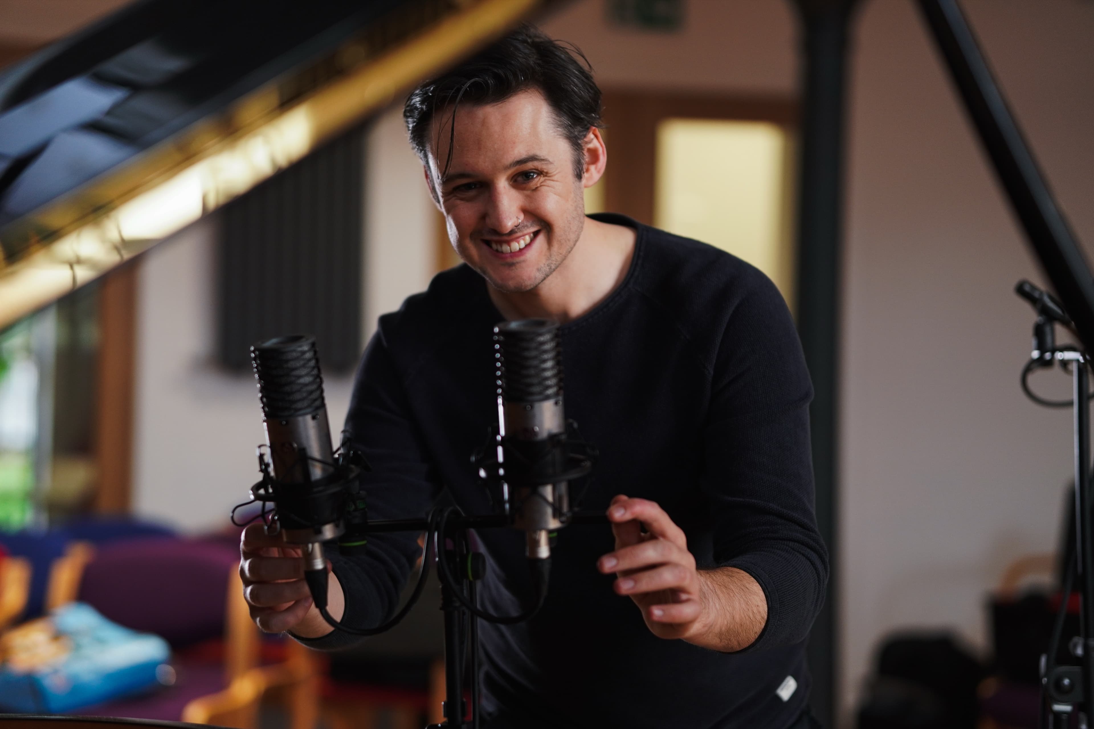 A smiling man adjusting studio microphones beside a grand piano