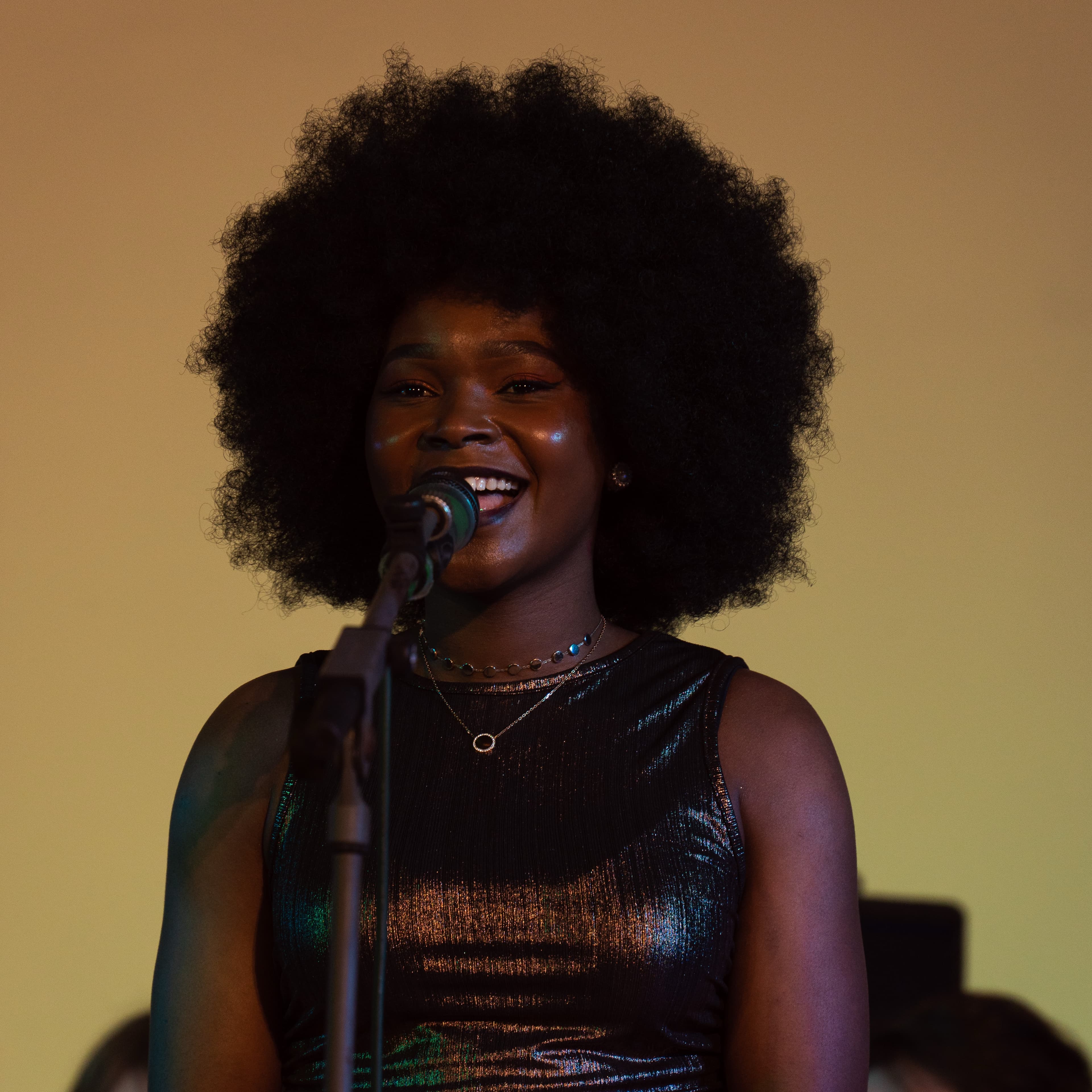 A singer smiling while performing into a microphone against a warm amber backdrop