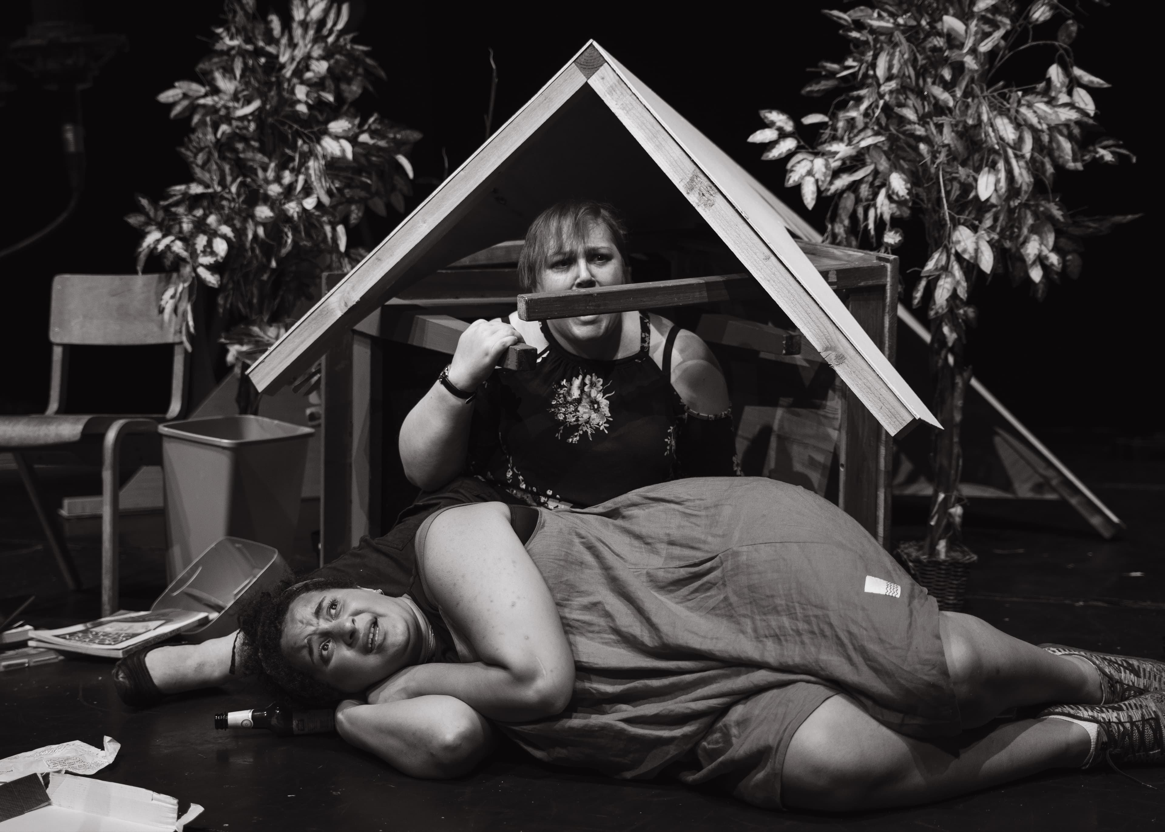 Two performers in a dramatic scene, one lying on the ground and the other crouching over her holding a wooden beam, black and white