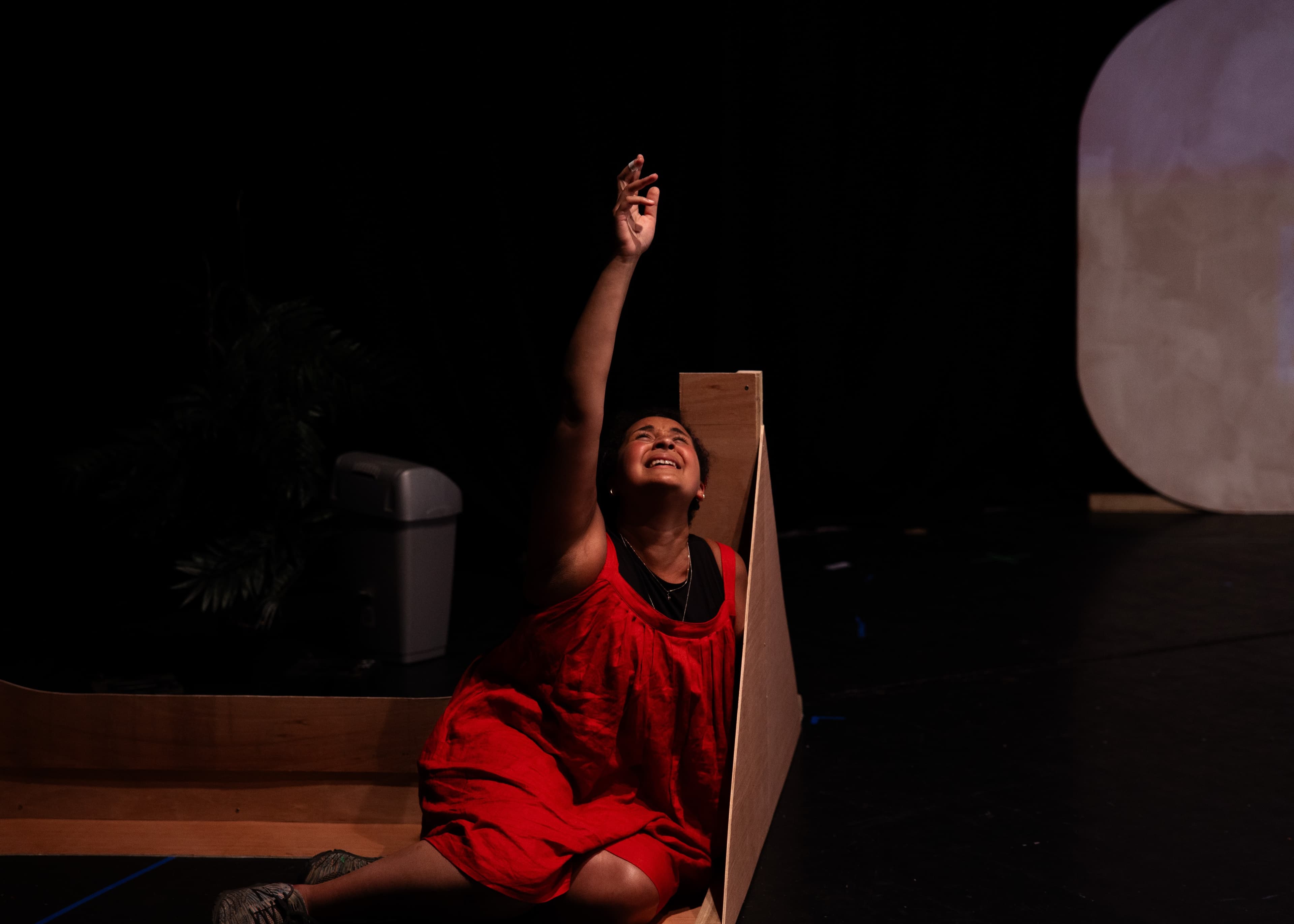 A performer in a red dress reaching upward while seated against a wooden ramp on a dark stage