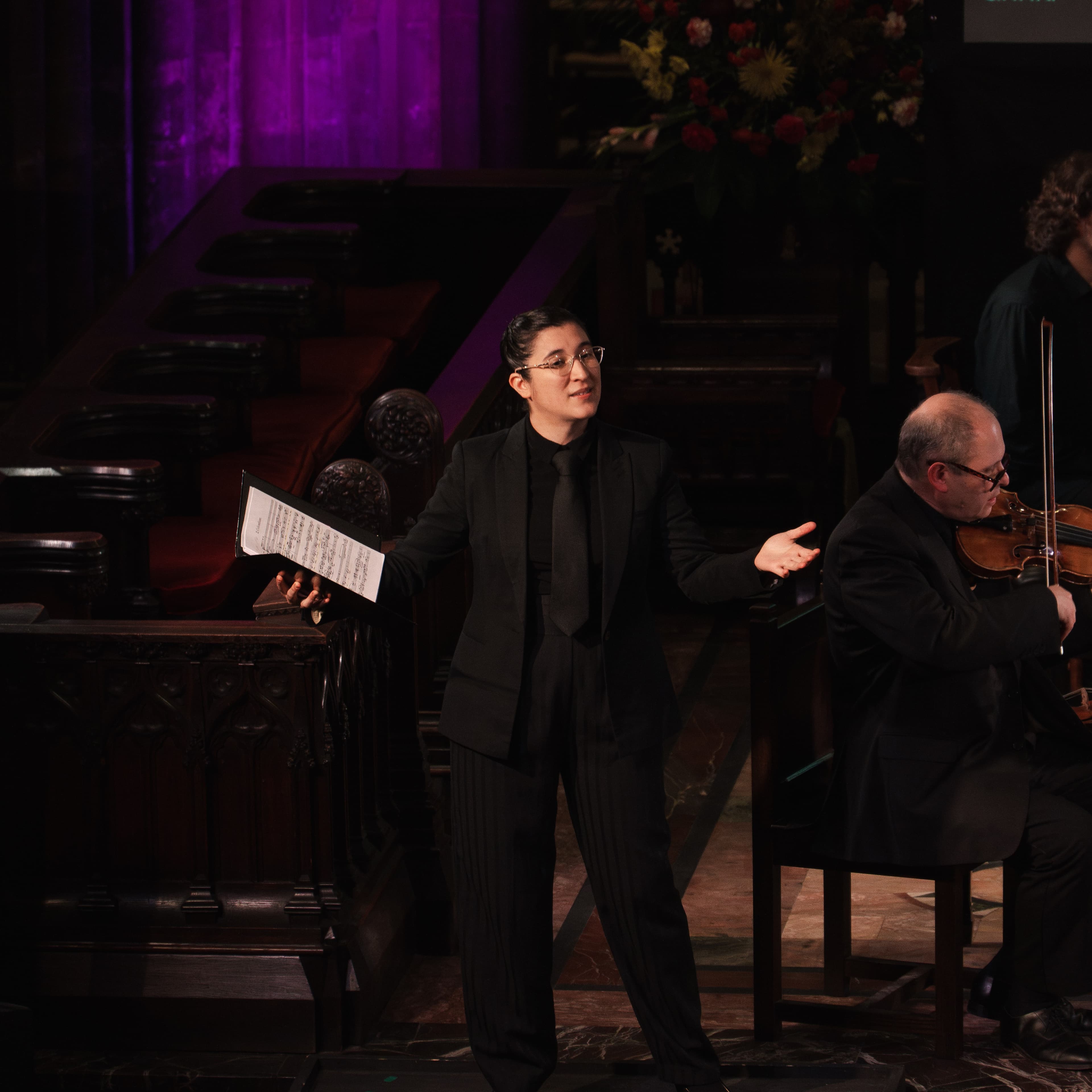 A performer in a suit and tie gesturing mid-performance while holding a music folder, with a violinist behind