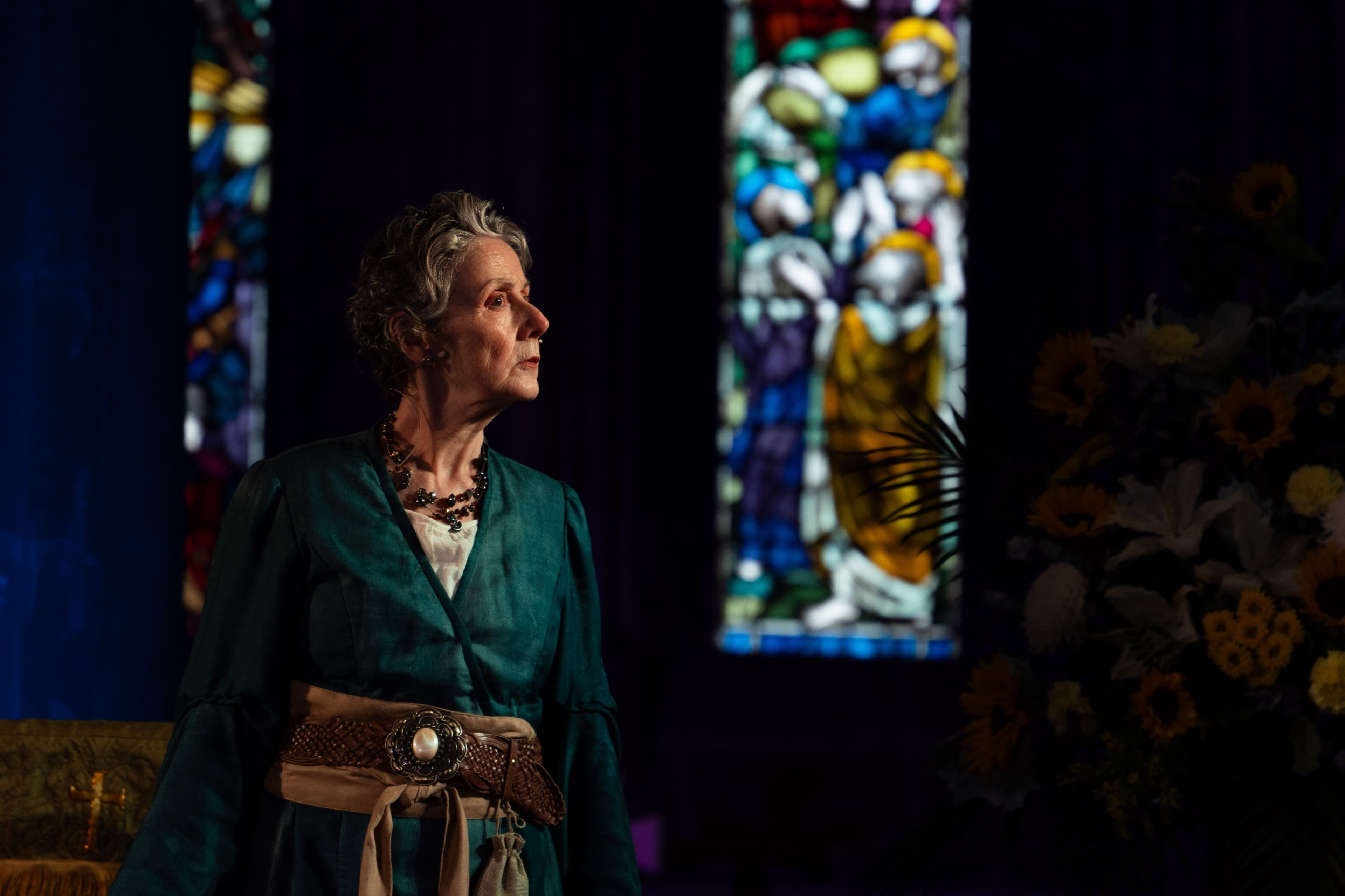 An older performer in costume standing in profile beside a stained glass window