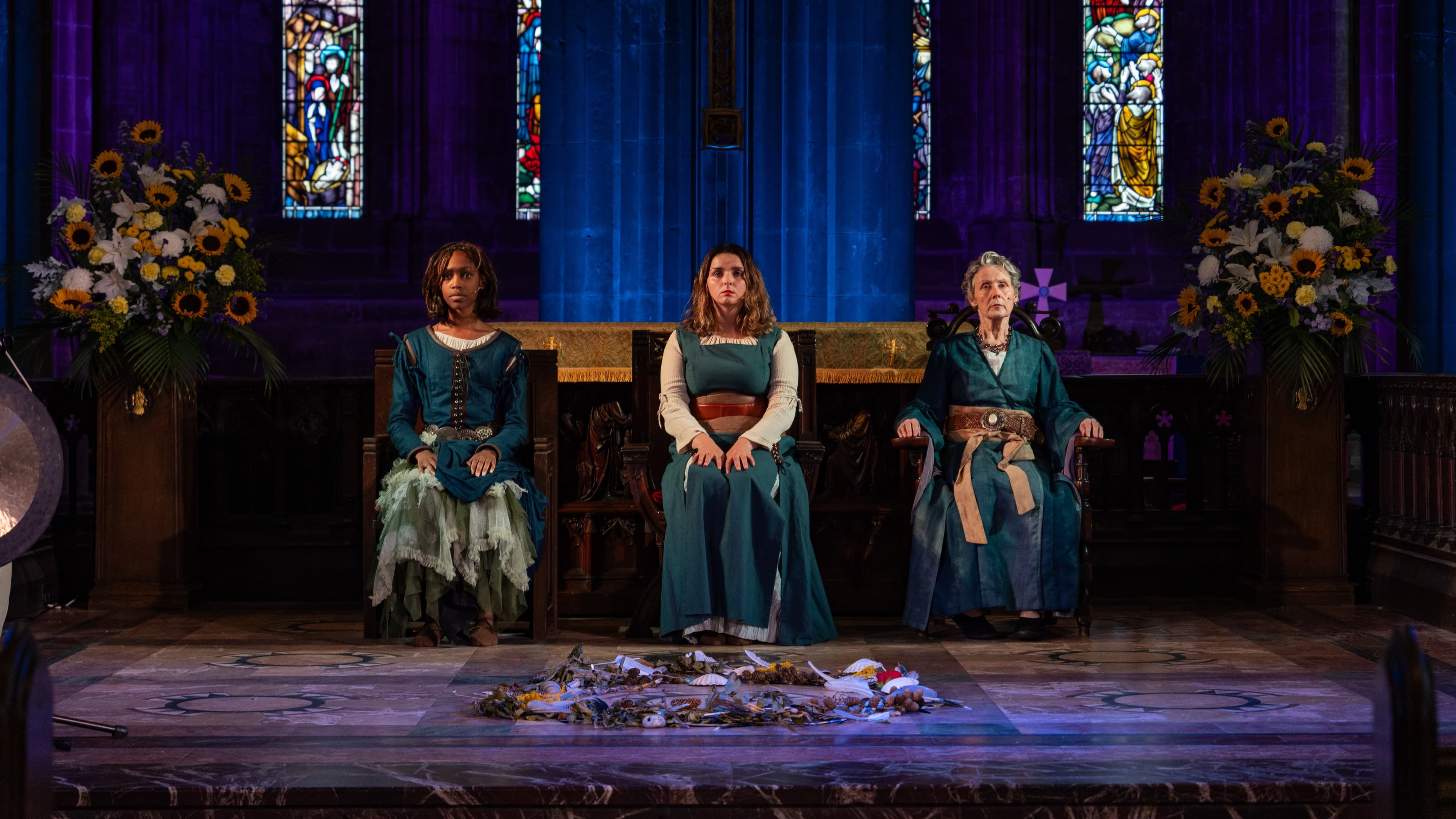 Three seated performers in costume on a church stage flanked by stained glass windows