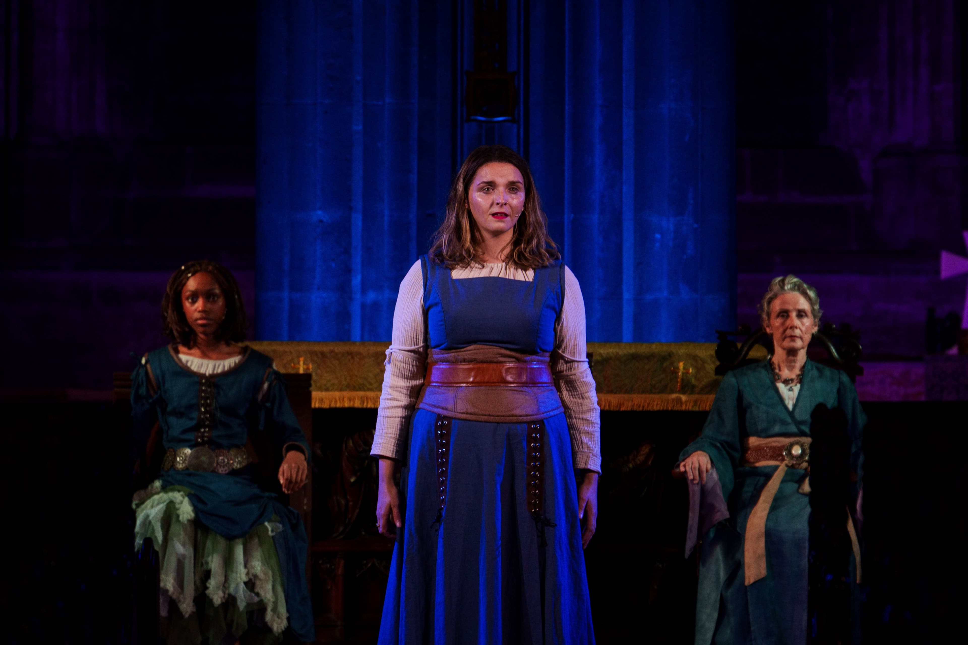Three performers in blue medieval-style costume standing on a stage lit with blue light