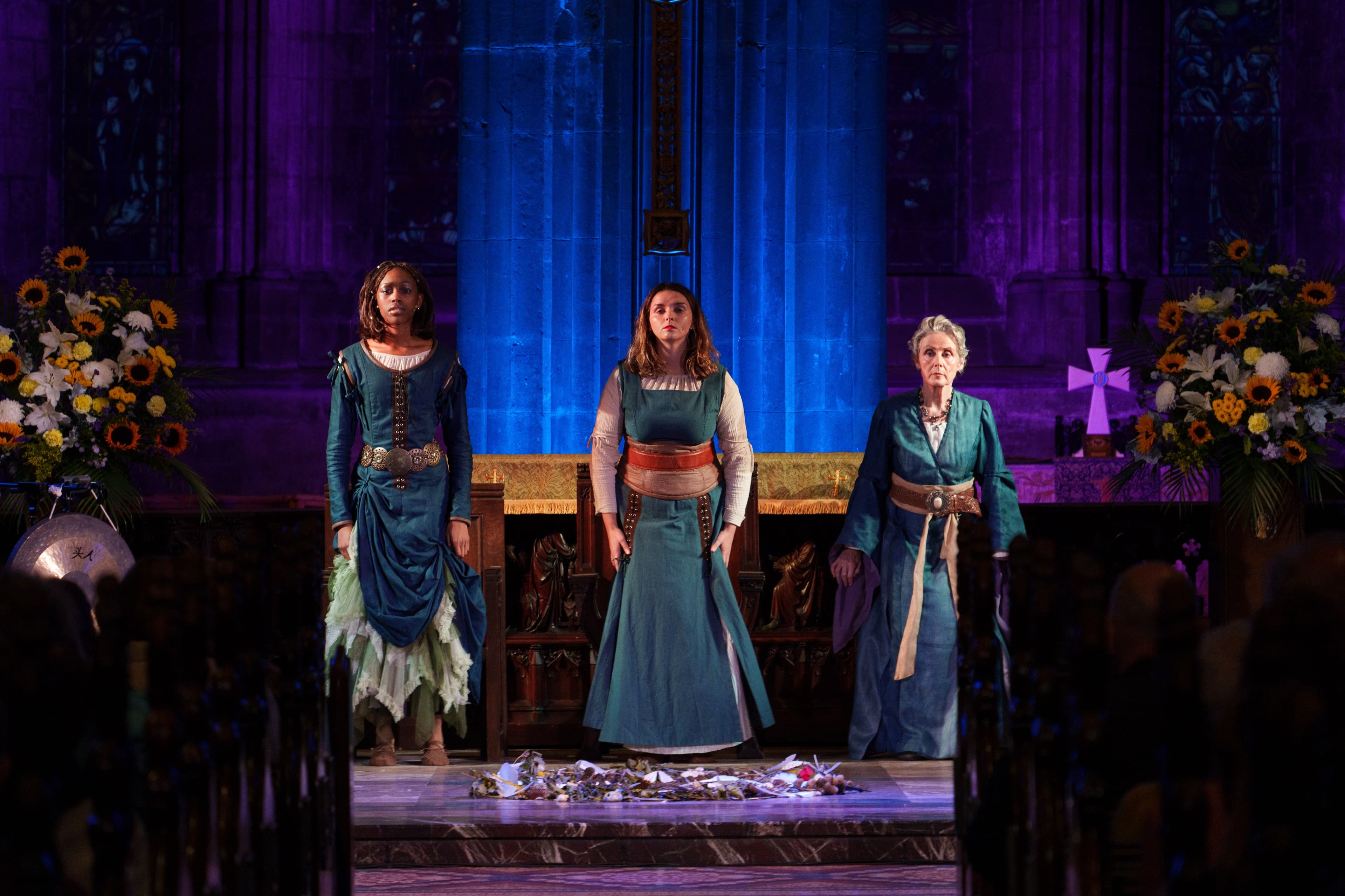 Three performers in costume seated on stage in a church setting with floral arrangements