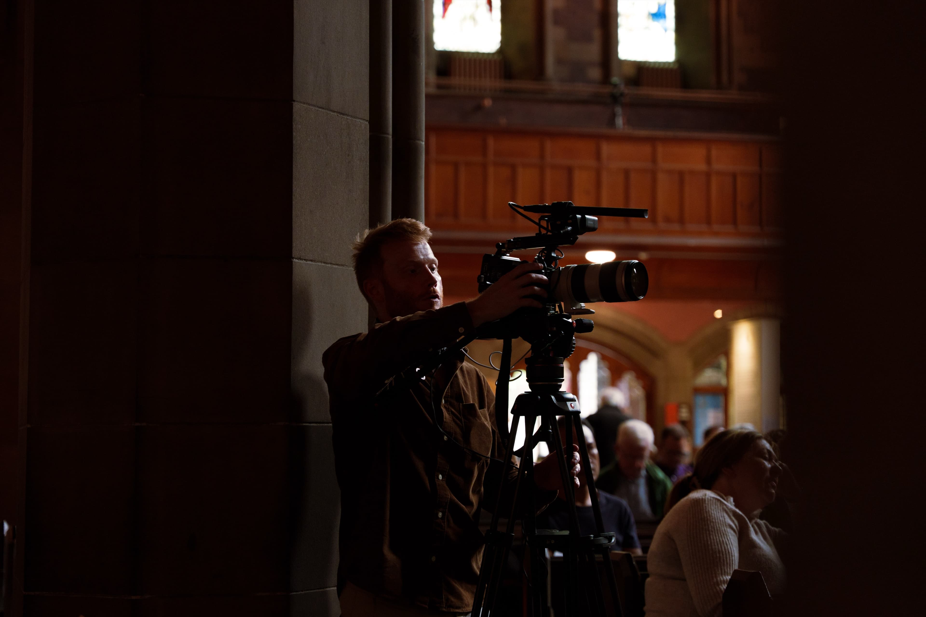 A videographer silhouetted operating a tripod-mounted camera with boom mic in a church interior