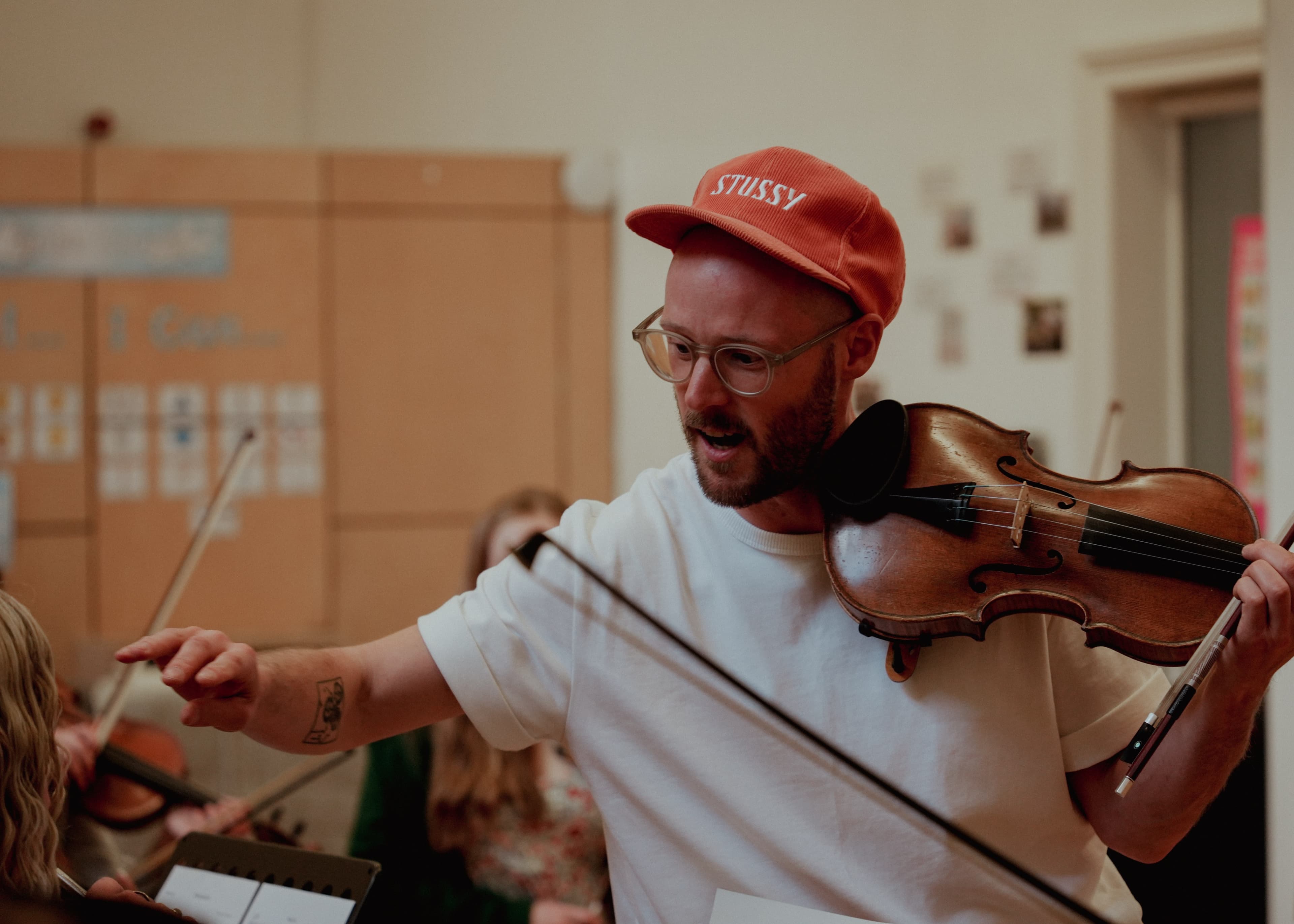A violinist in a cap pointing while playing and calling out during a rehearsal