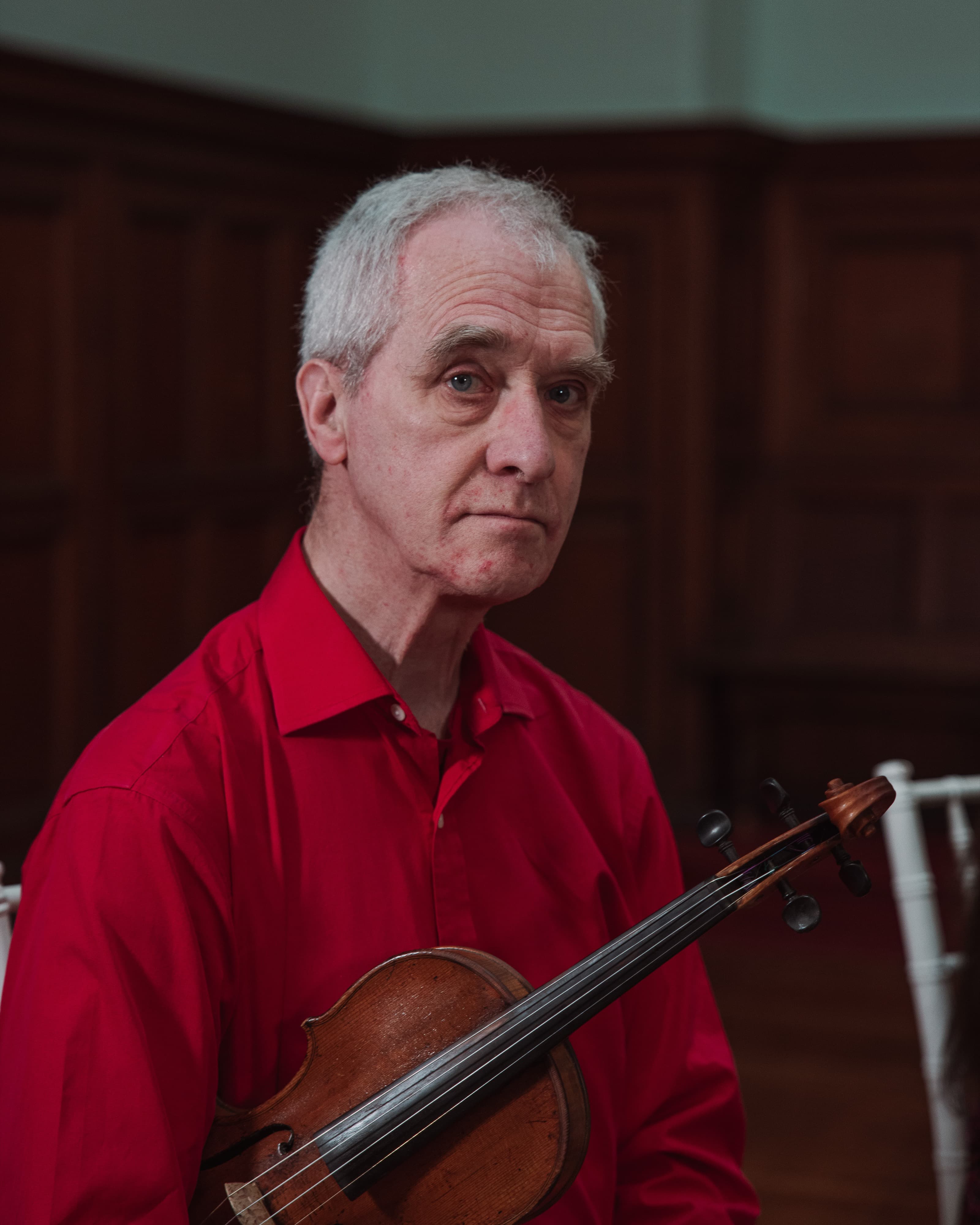 A portrait of a man in a red shirt holding a violin in a wood-panelled room