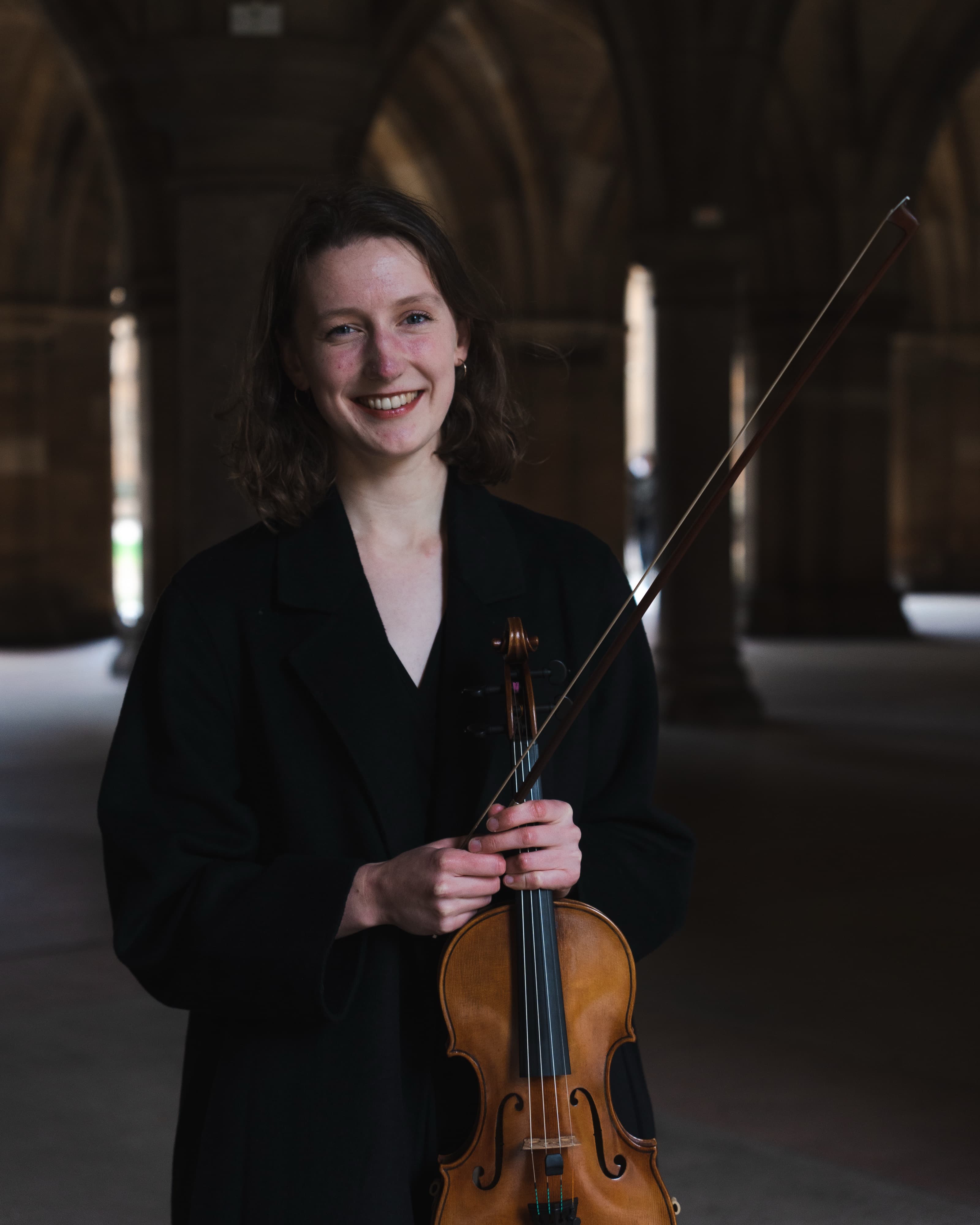 A portrait of a woman in a black coat smiling while holding a violin and bow in a stone-arched cloister