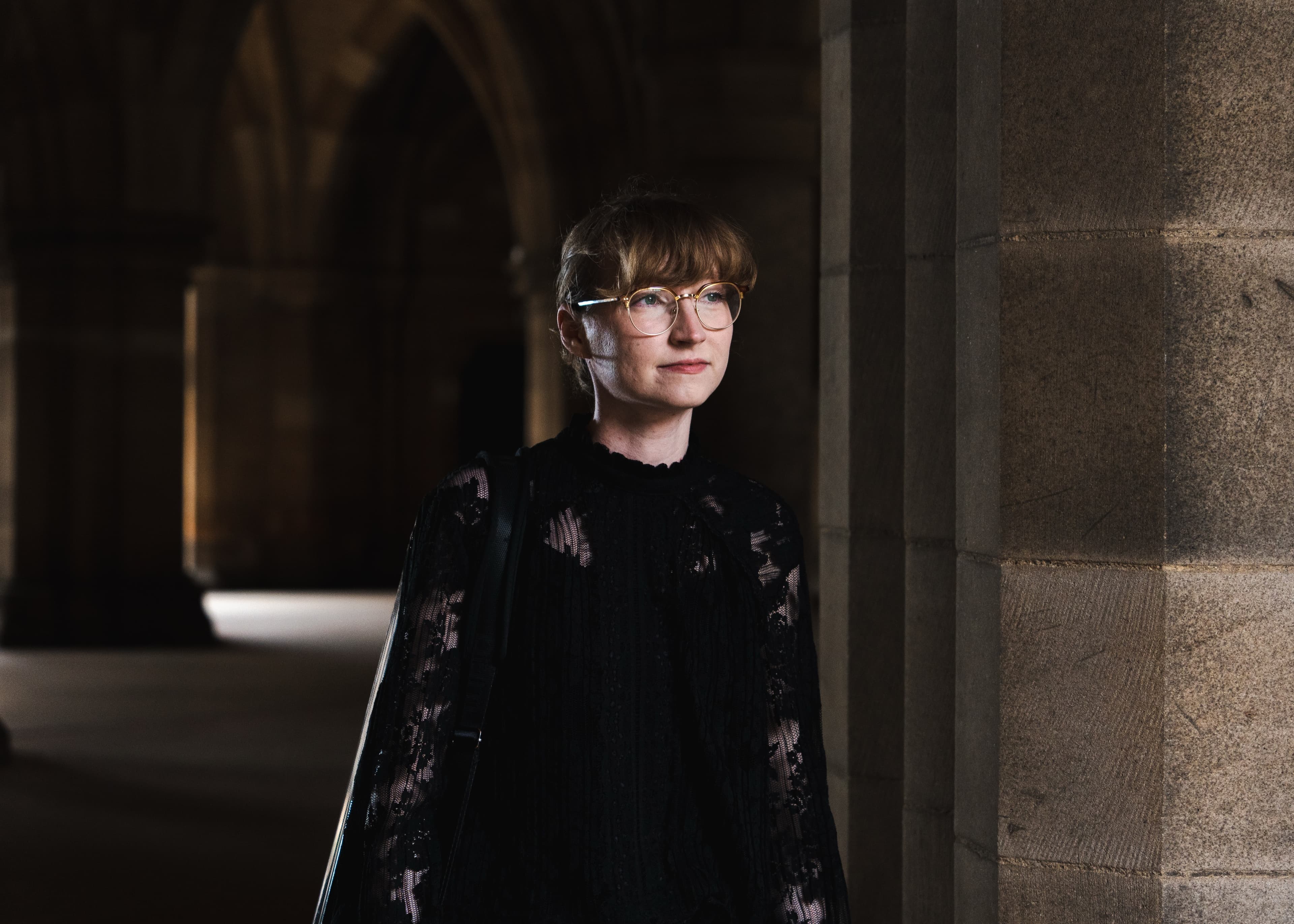 A portrait of a woman in glasses and a black lace top standing in a stone archway, looking up and to the side