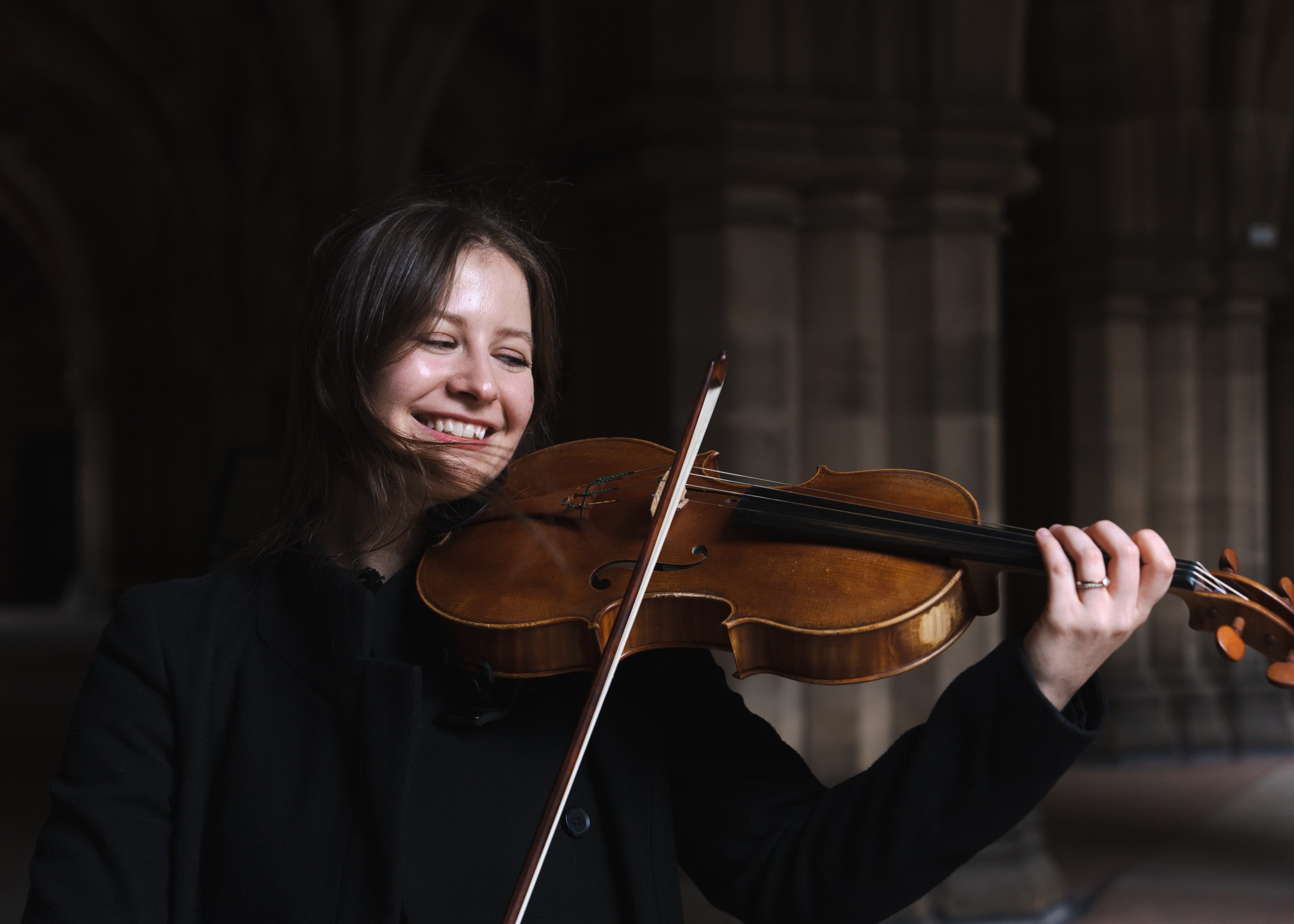 A portrait of a woman smiling while playing violin in a stone-arched cloister