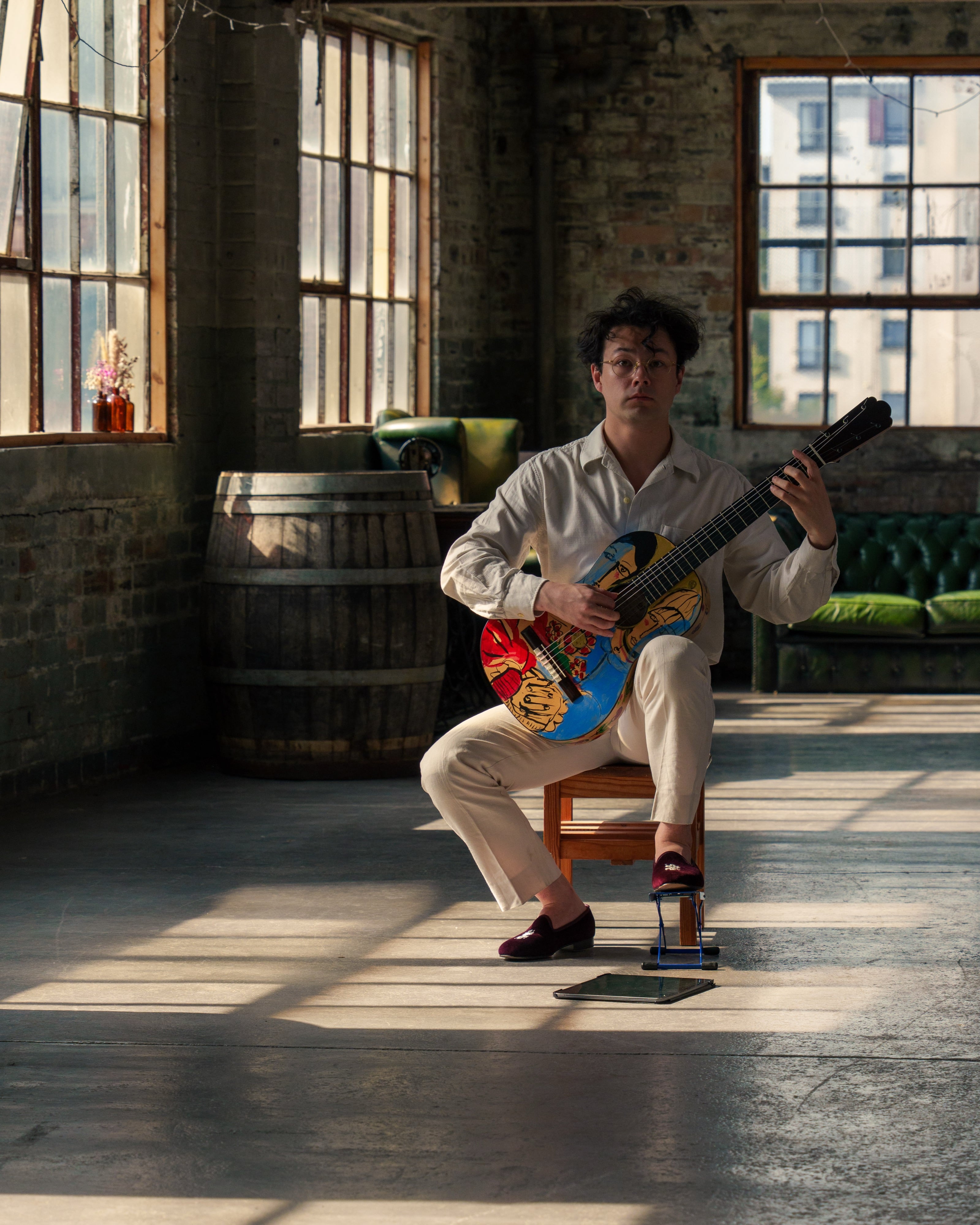 A guitarist looking directly at the camera, holding a hand-painted classical guitar in a sunlit industrial space